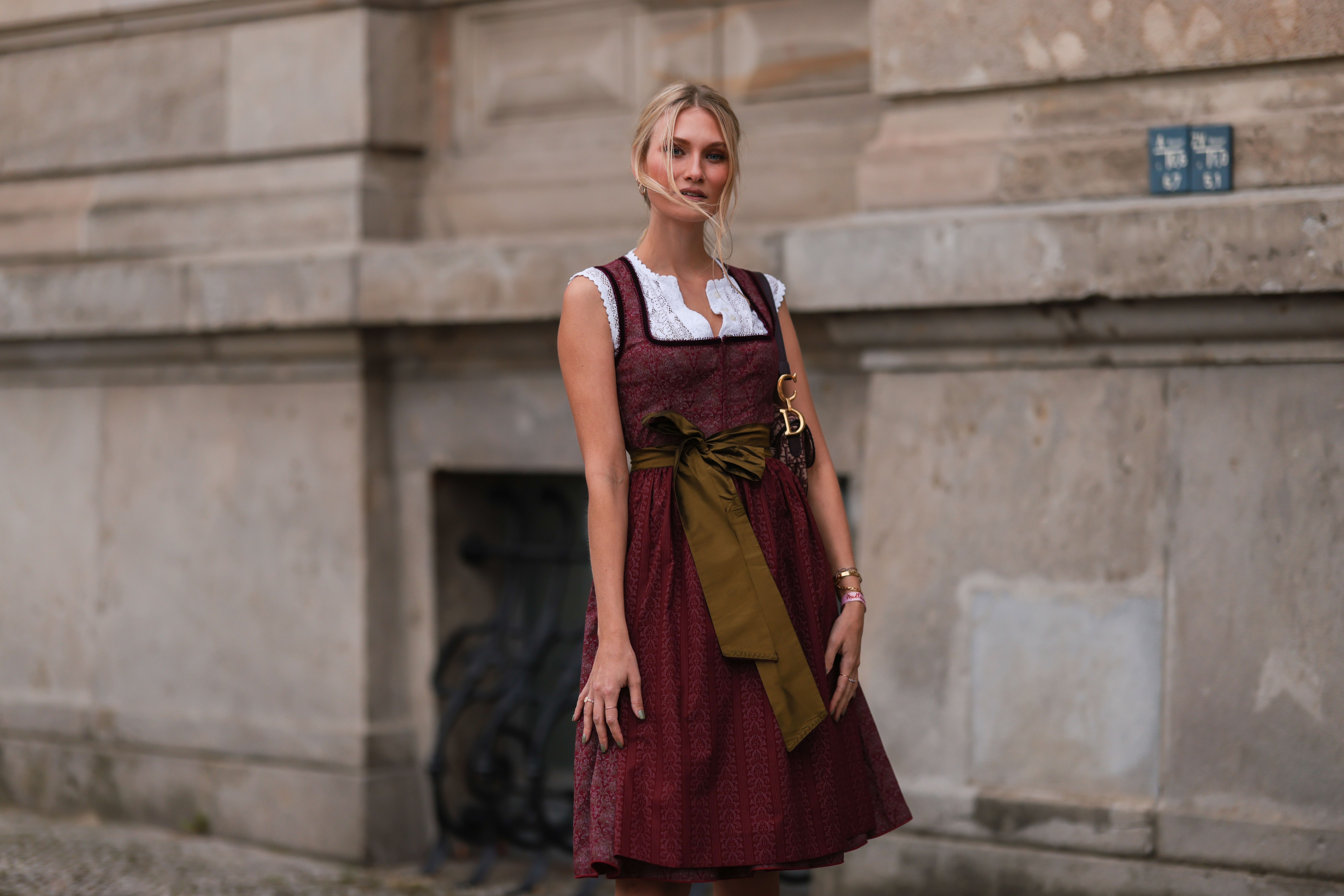 Frau in rotem Dirndl mit weißer Bluse und olivgrüner Schleife steht vor einer Steinmauer.