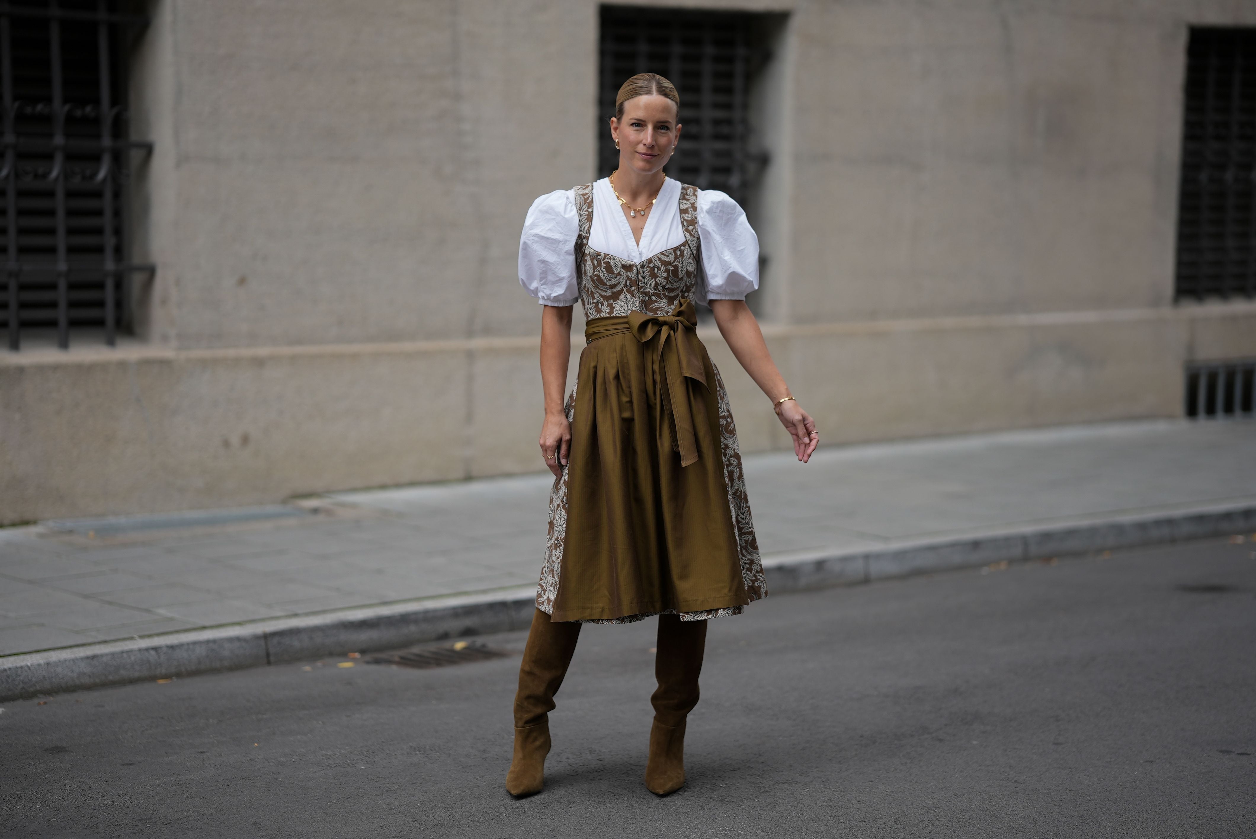 Frau in traditioneller Tracht mit weißer Bluse, braunem Dirndl, Schürze und braunen Stiefeln auf der Straße.