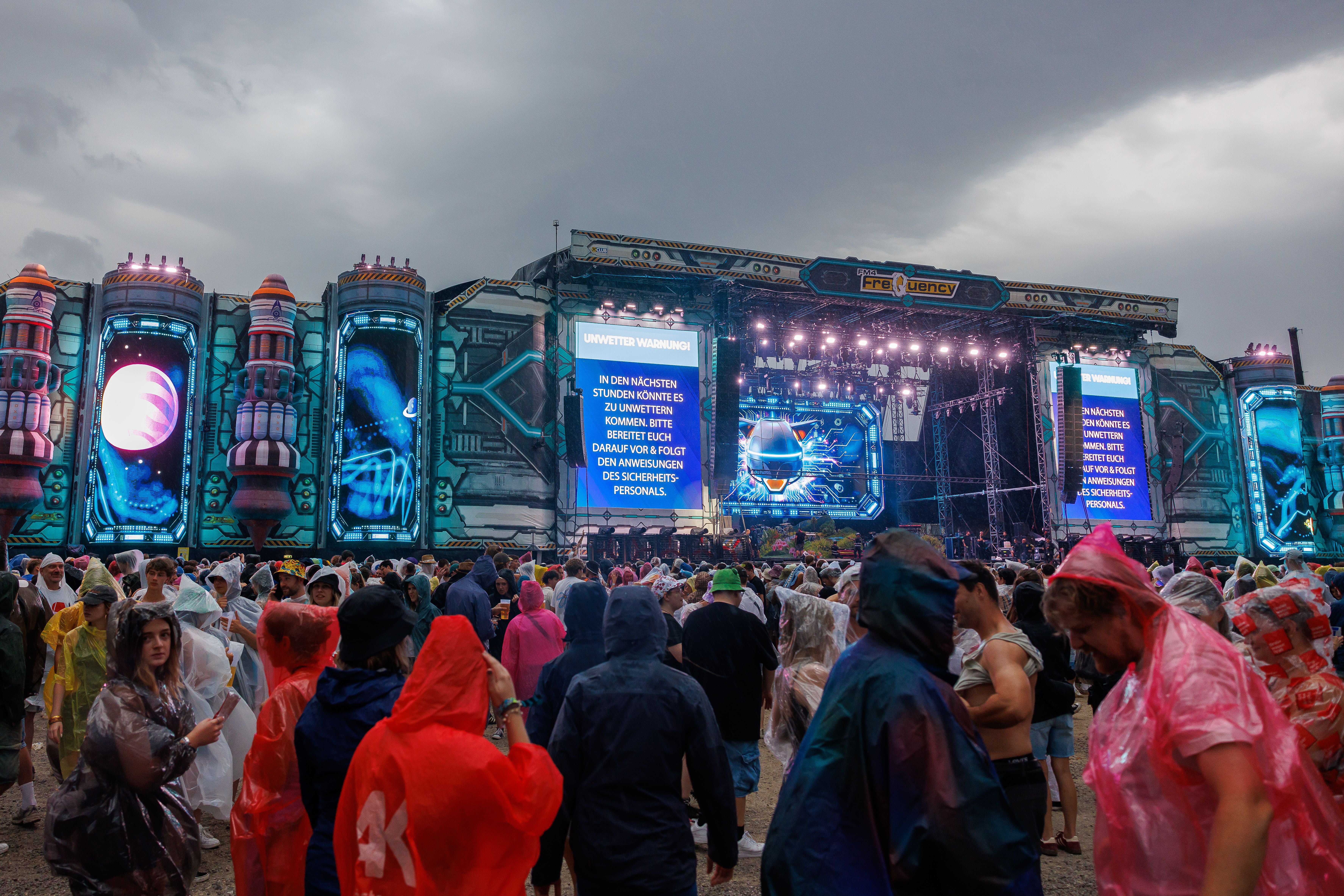 Menschen in Regenponchos stehen vor der Space Stage beim Frequency Festival mit Unwetterwarnung auf Monitoren.