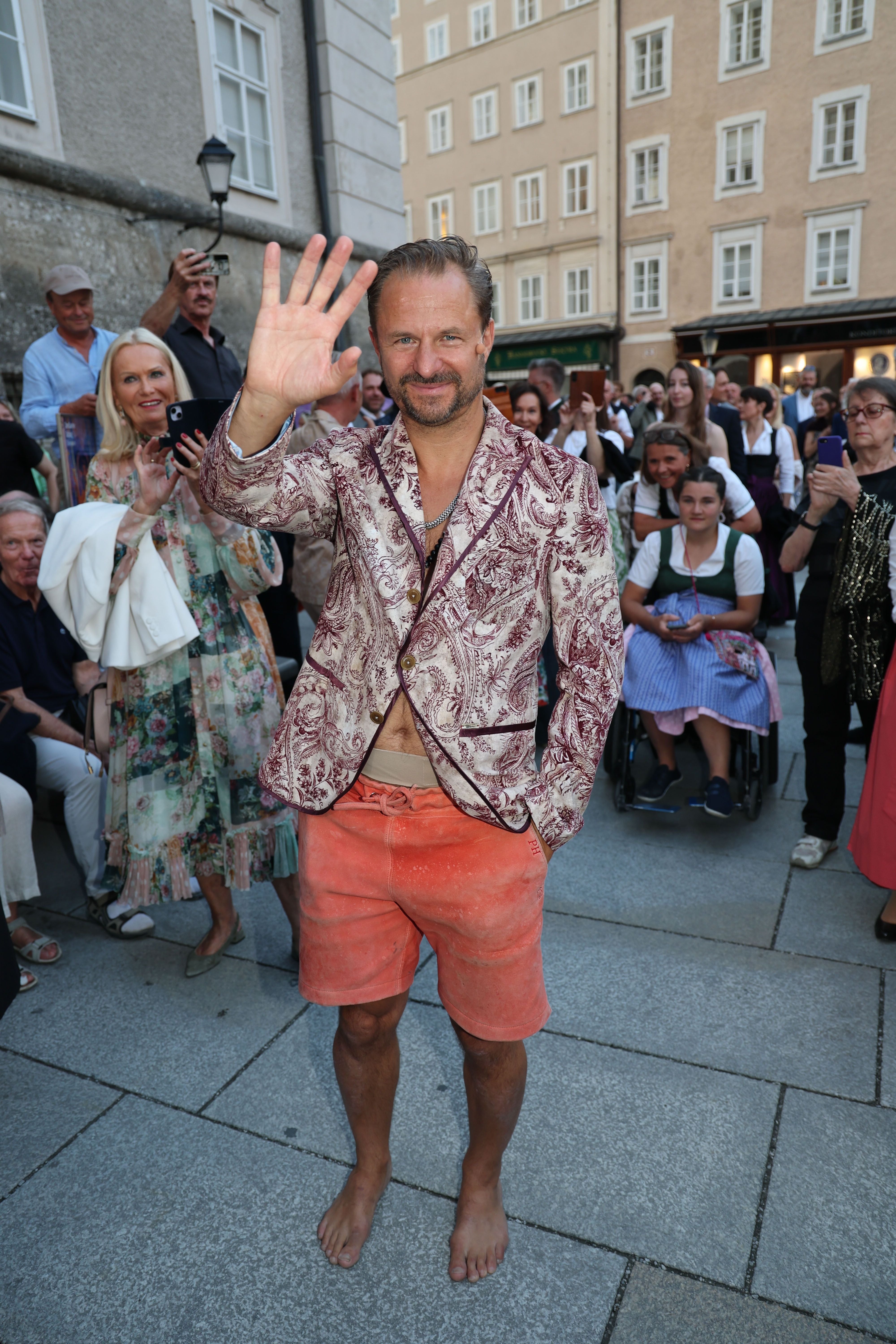 Philipp Hochmair läuft barfuß in bunter Hose und gemustertem Sakko durch eine Menschenmenge.