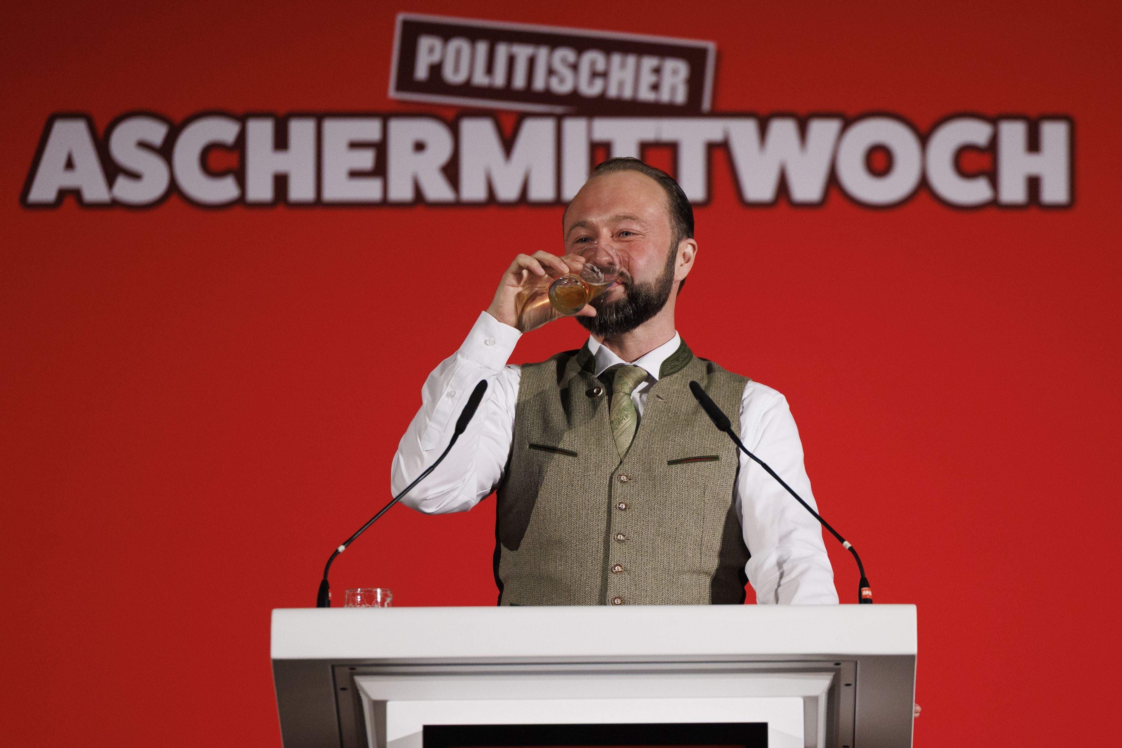 Ein Mann trinkt an einem Rednerpult beim politischen Aschermittwoch vor rotem Hintergrund.