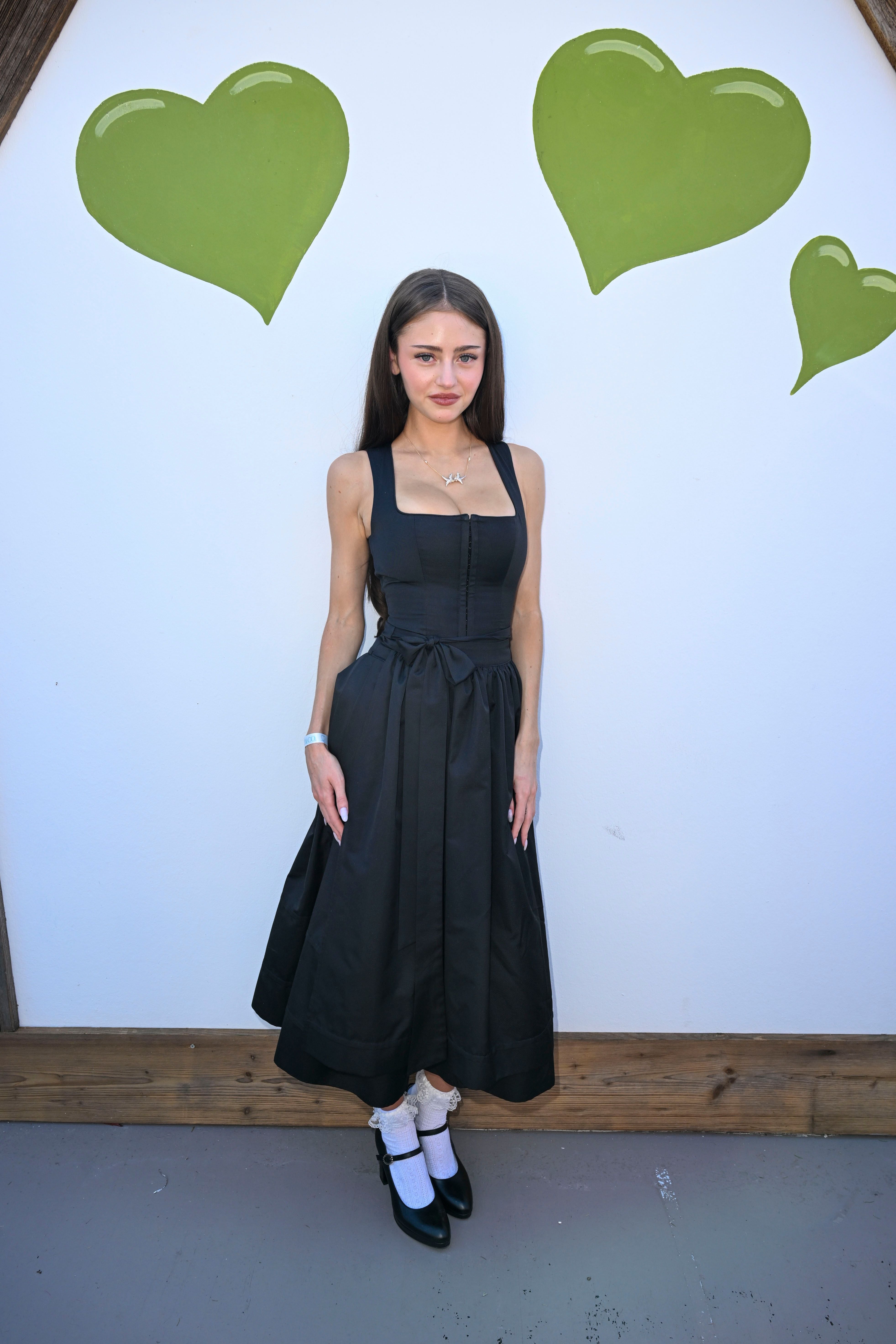 Frau in schwarzem Kleid steht vor weißer Wand mit grünen Herzmotiven.