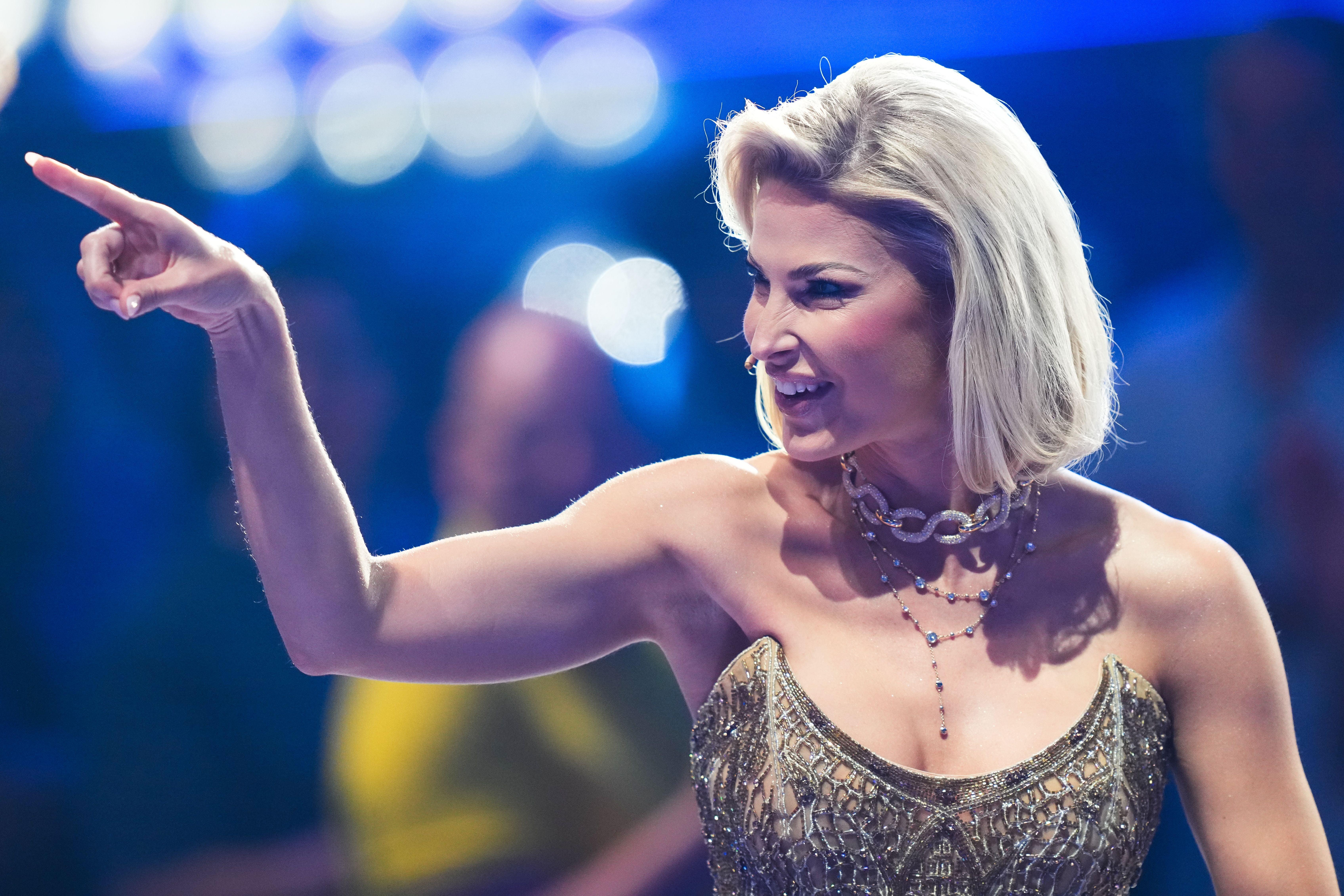 Eine Frau mit blondem Haar und Glitzerkleid zeigt lächelnd mit dem Finger nach links.