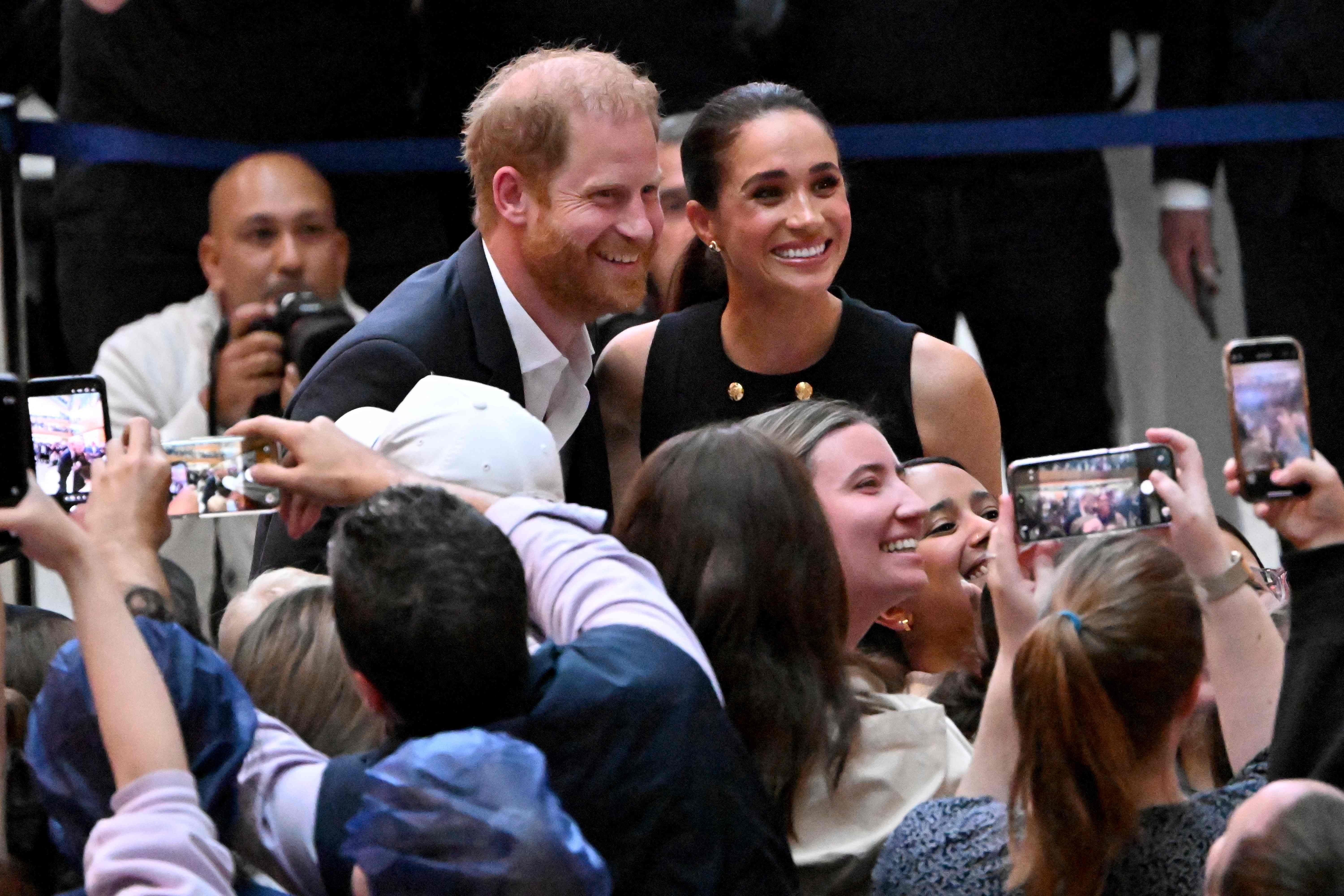 Prinz Harry und Meghan lächeln für Fotos mit einer Menschenmenge, viele halten Handys hoch.