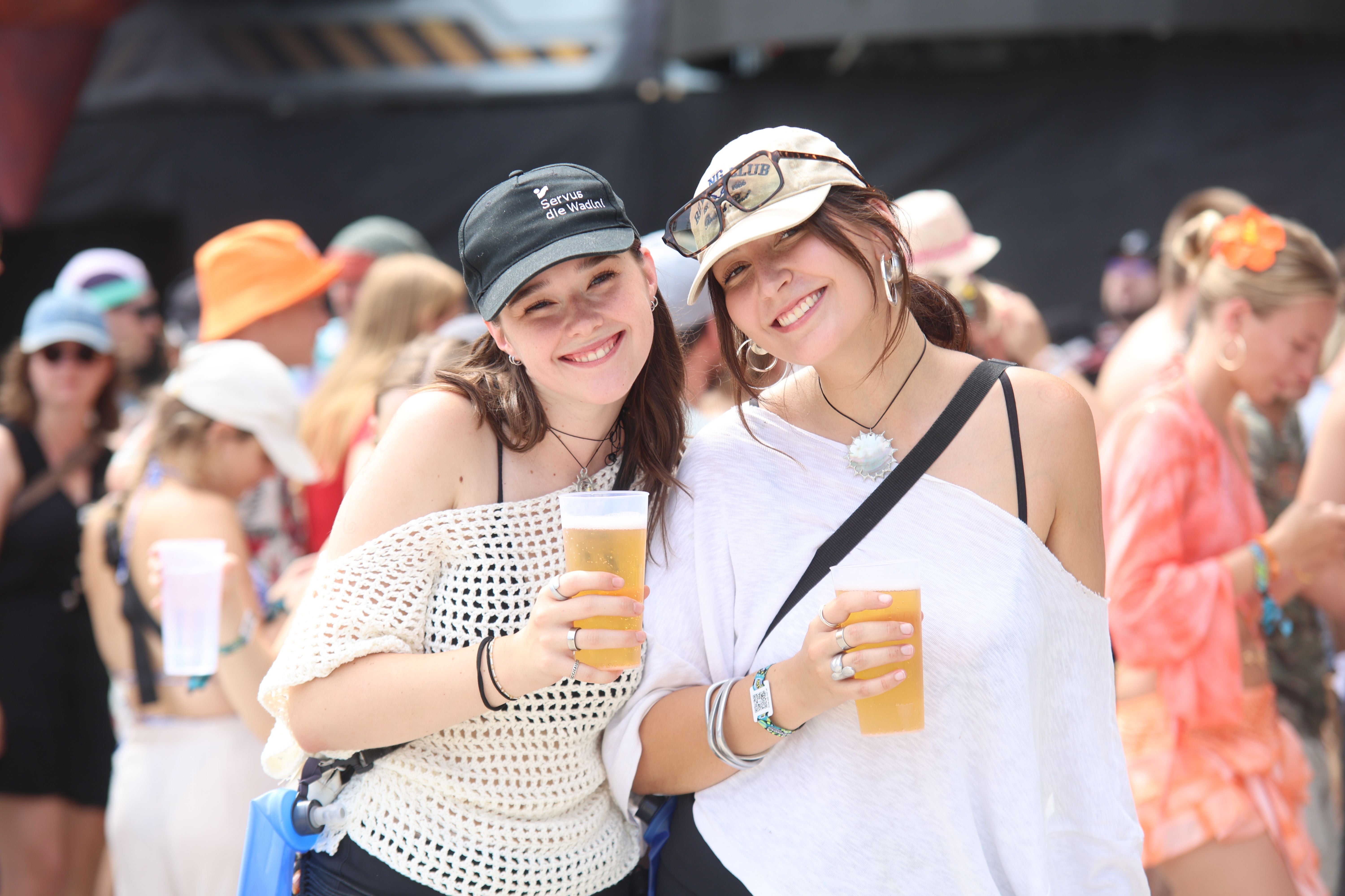 Zwei lächelnde junge Frauen mit Getränken auf einem Outdoor-Festival bei Sonnenschein.