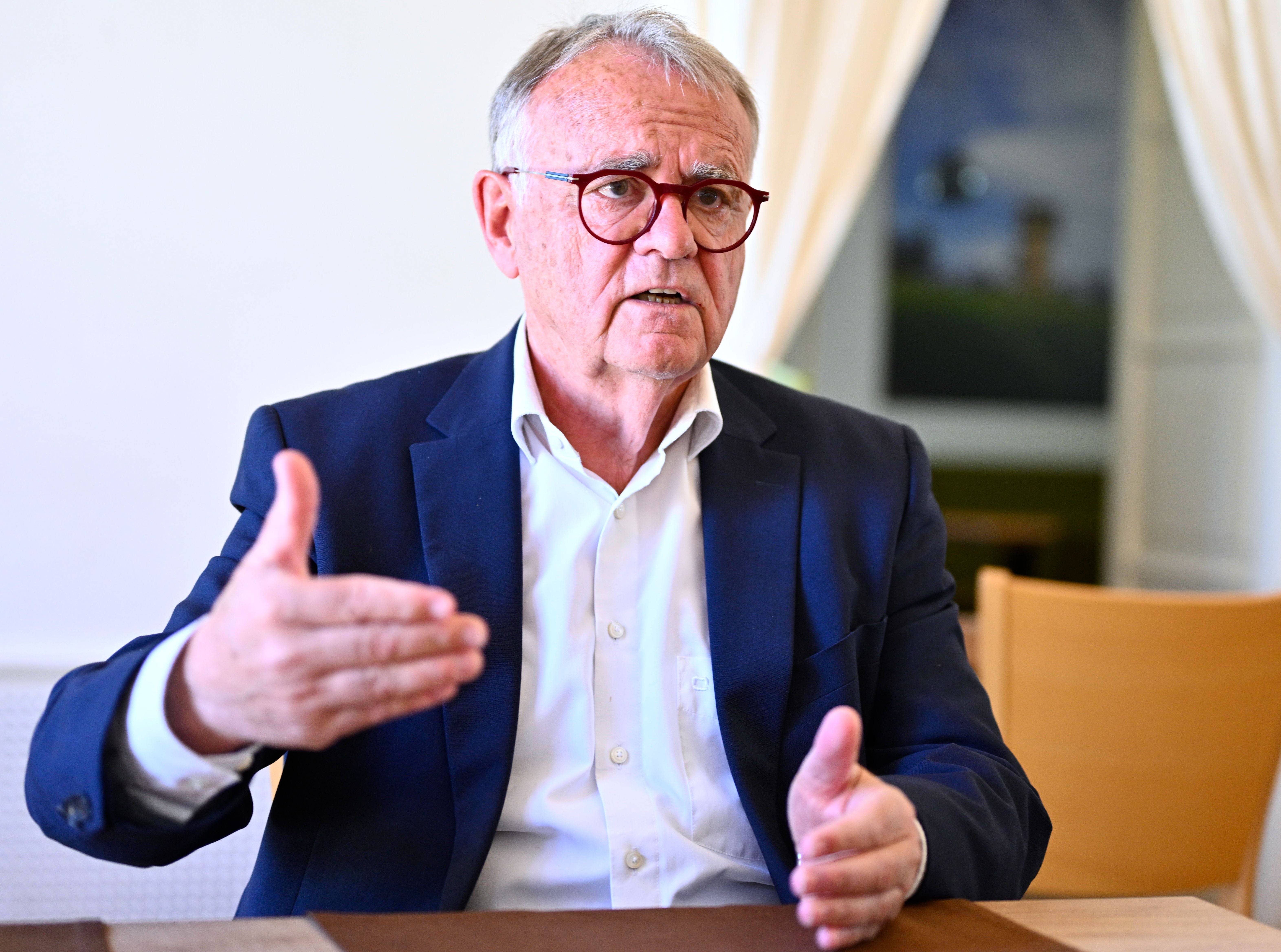 Ein älterer Mann im Anzug spricht gestikulierend in einem Interview in einem hellen Raum.