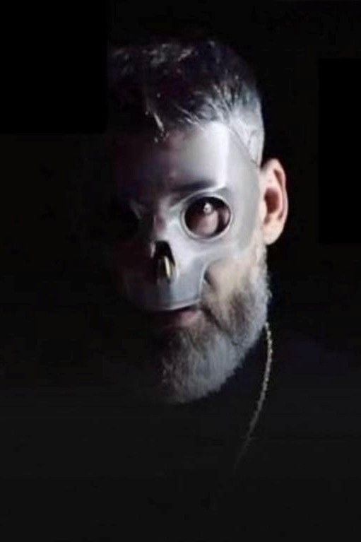 Ein Mann mit silberner Totenkopfmaske blickt in die Kamera, dunkler Hintergrund.