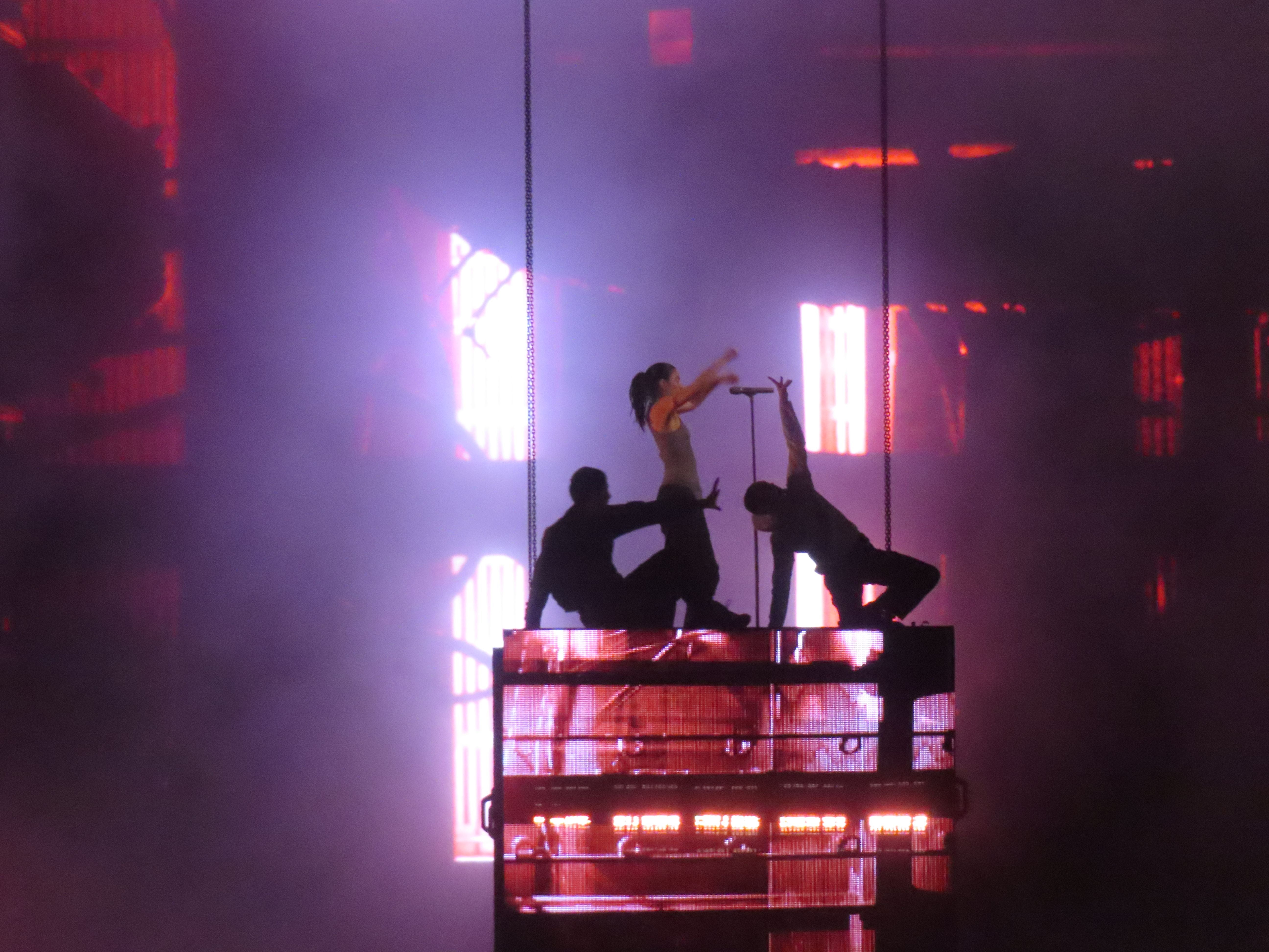 Drei Personen performen auf einer Bühne mit Lichteffekten und Nebel im Hintergrund.