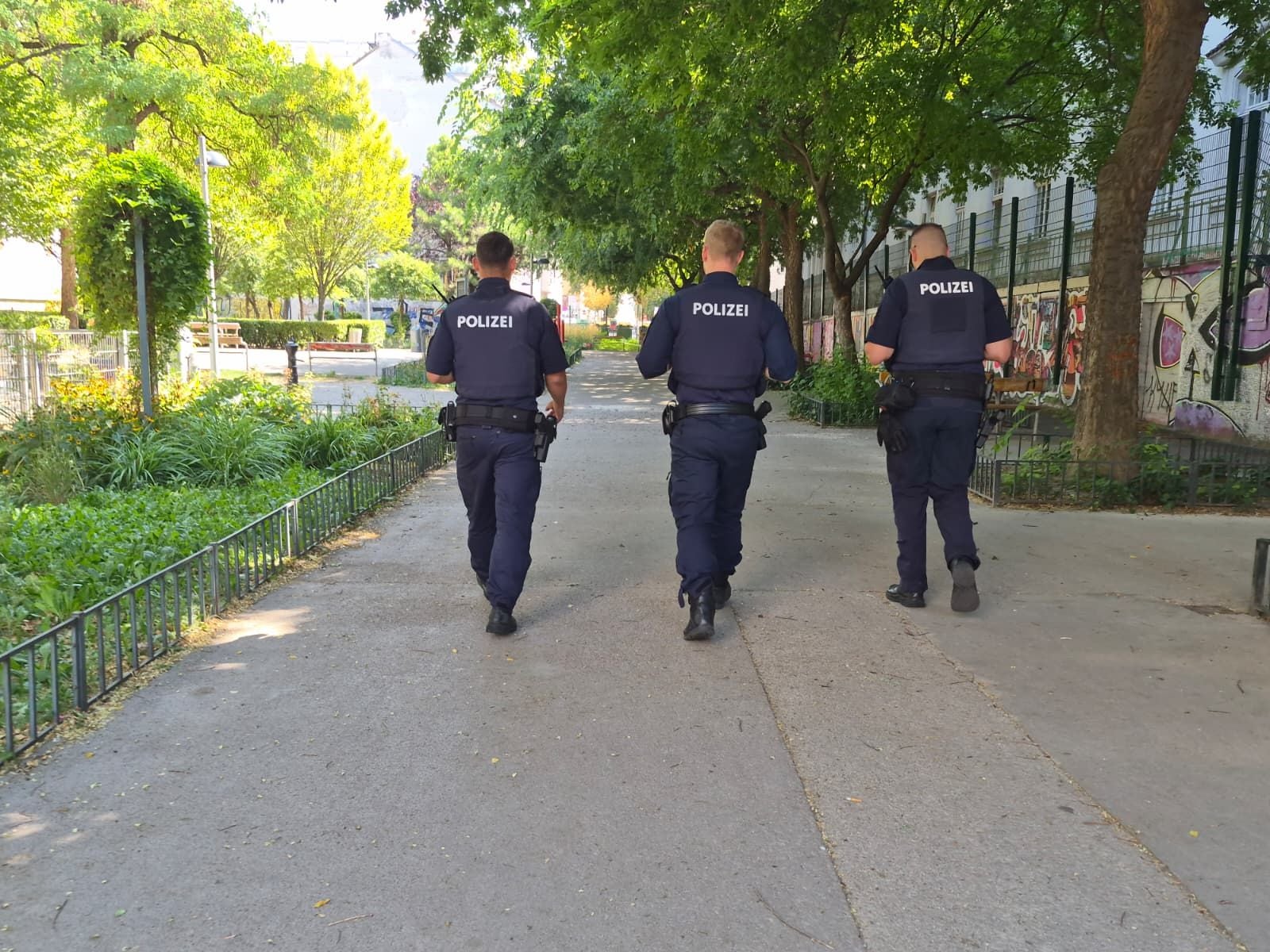 Drei Polizisten gehen in Uniform durch einen sonnigen Park mit viel Grün und Graffiti an einer Mauer.
