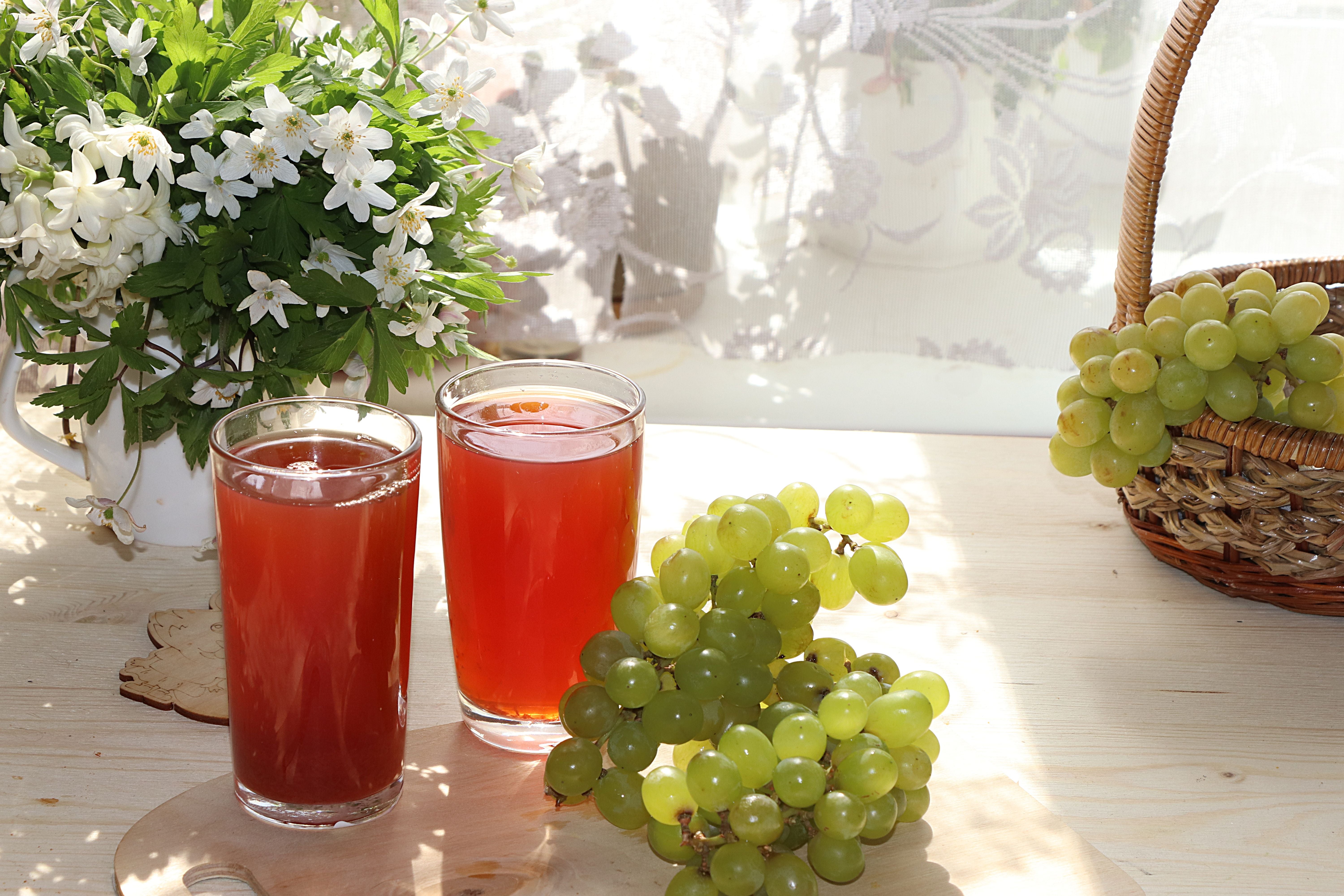 Zwei Gläser Traubensaft, grüne Weintrauben und ein Blumengesteck auf einem sonnigen Holztisch.