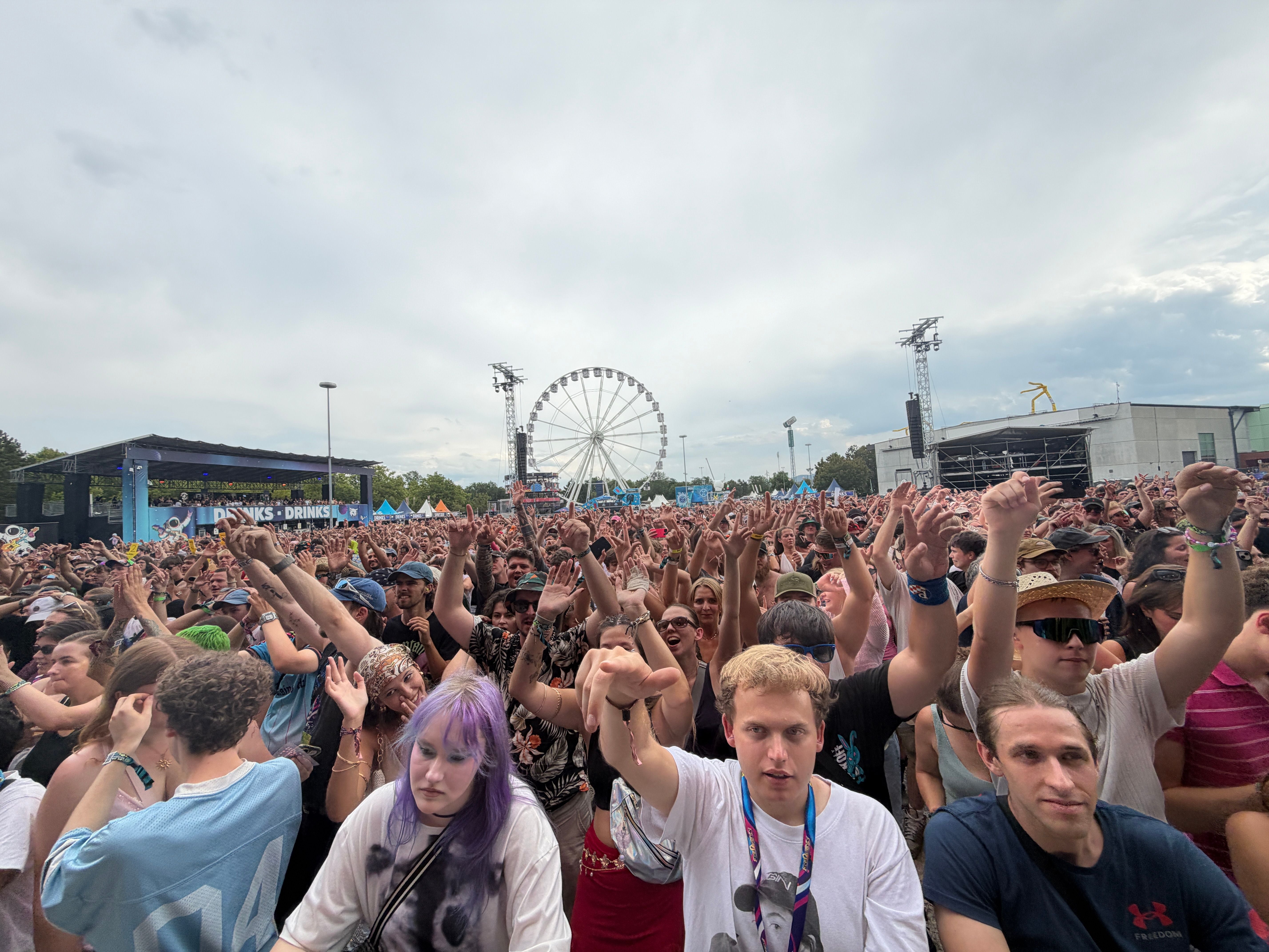 Große Menschenmenge feiert vor einer Festivalbühne, im Hintergrund ein Riesenrad.