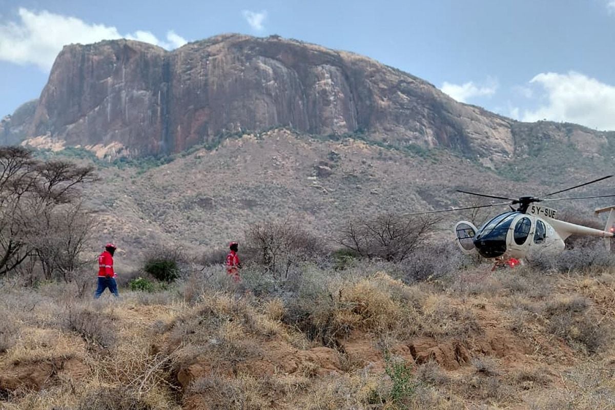 Zwei Rettungskräfte nähern sich einem abgestürzten Hubschrauber in einer bergigen, trockenen Landschaft.
