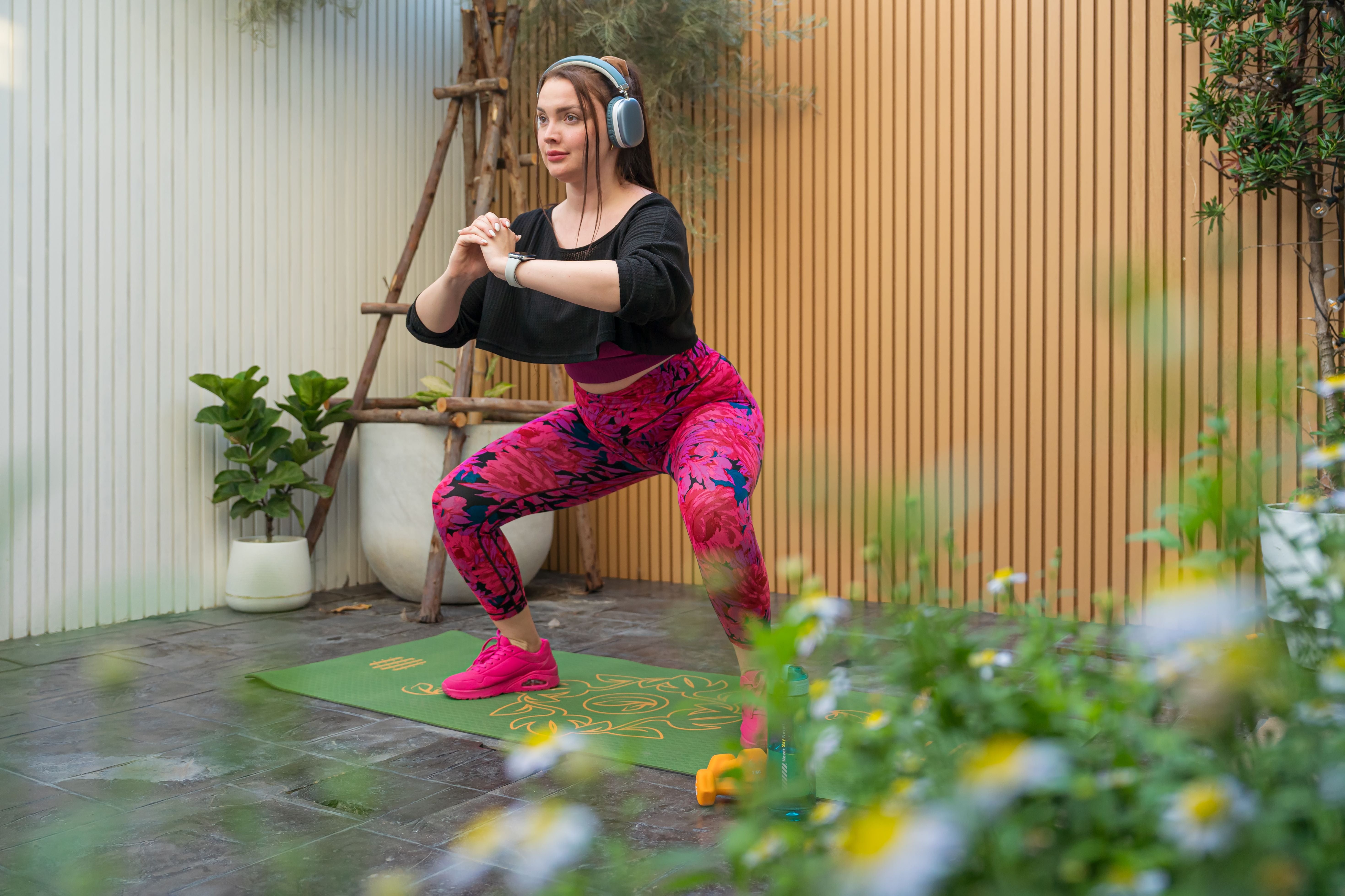 Junge Frau macht mit Kopfhörern Kniebeugen auf einer Matte in einem Garten.