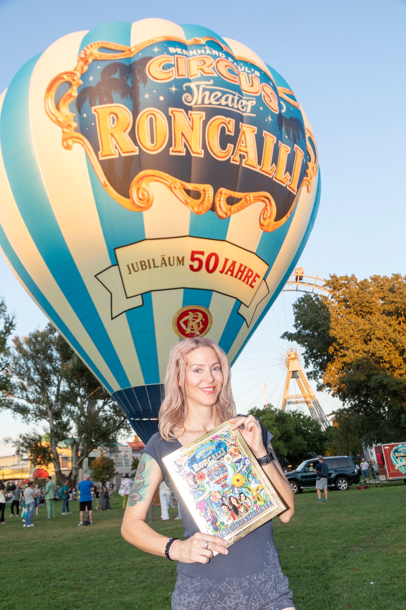Frau hält Plakat vor Roncalli-Heißluftballon zum 50-jährigen Jubiläum in einem Park in Wien.