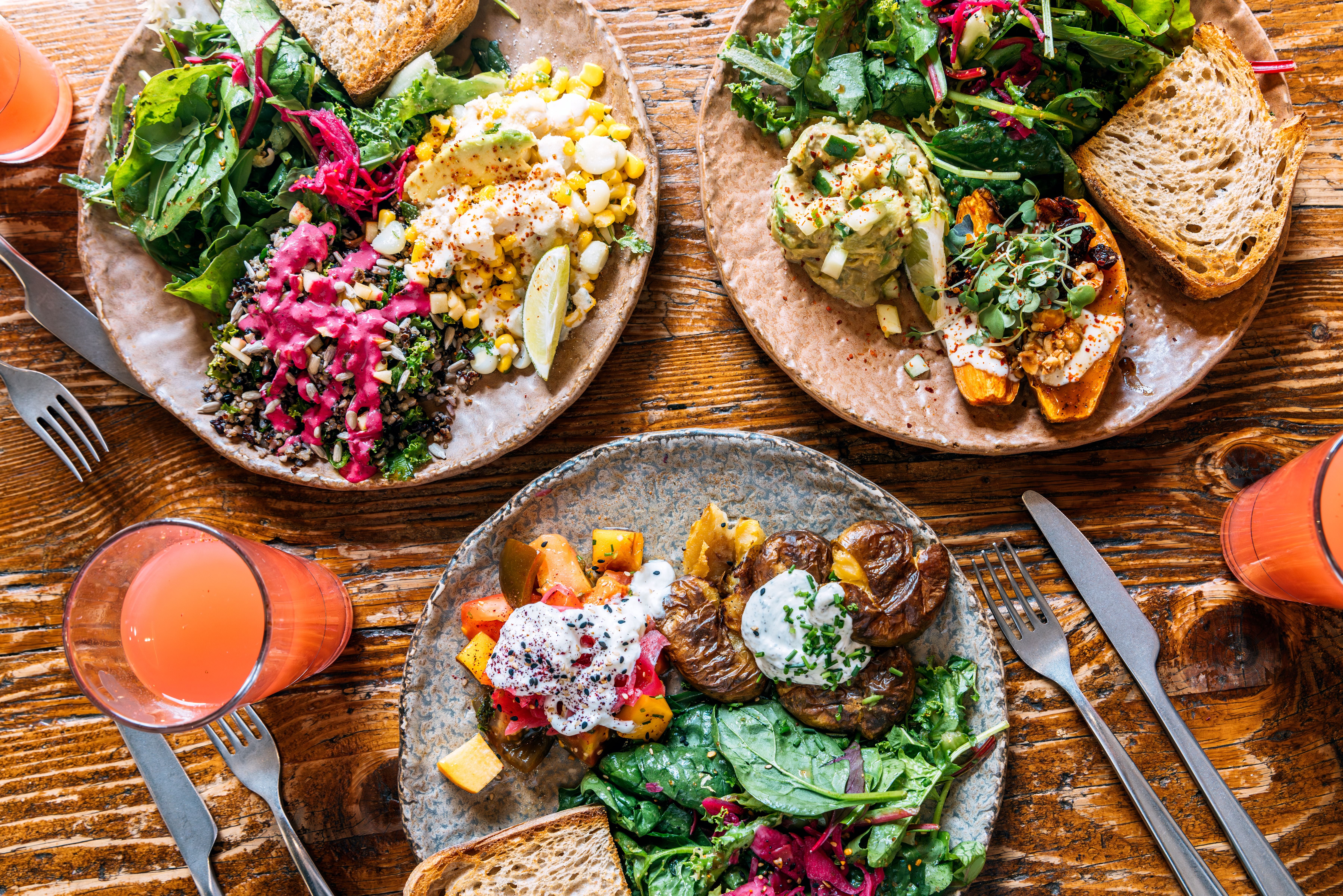 Drei Teller mit vegetarischen Gerichten, frischem Salat, Brot und zwei Gläsern Saft auf Holztisch.