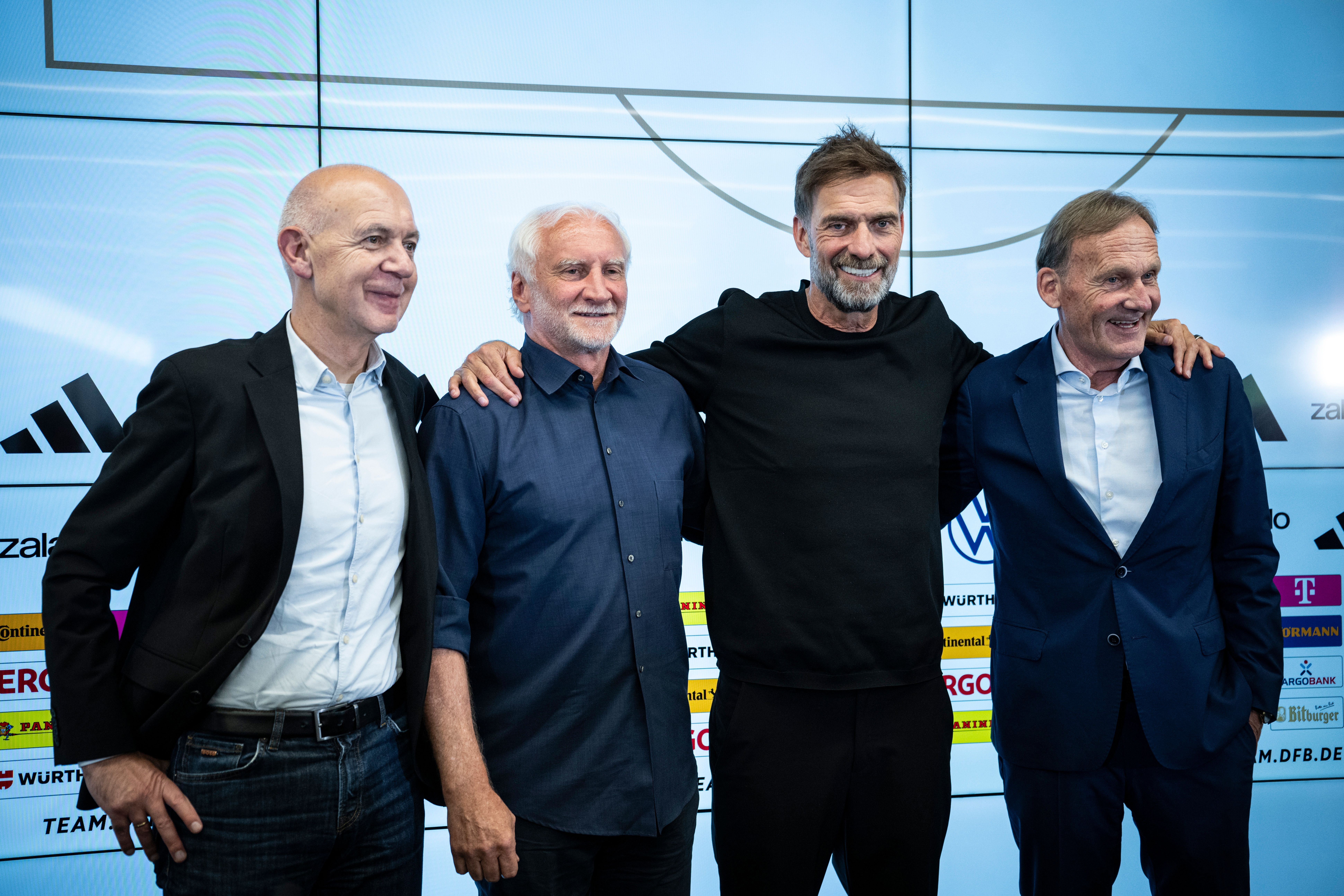 Vier Männer stehen lächelnd nebeneinander vor einer Sponsorenwand bei einer Pressekonferenz.