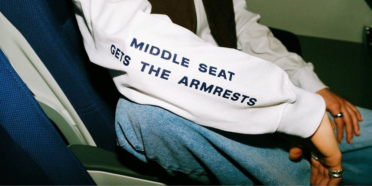 Person im Flugzeug mit Pullover, auf dem „Middle Seat Gets The Armrests“ steht.