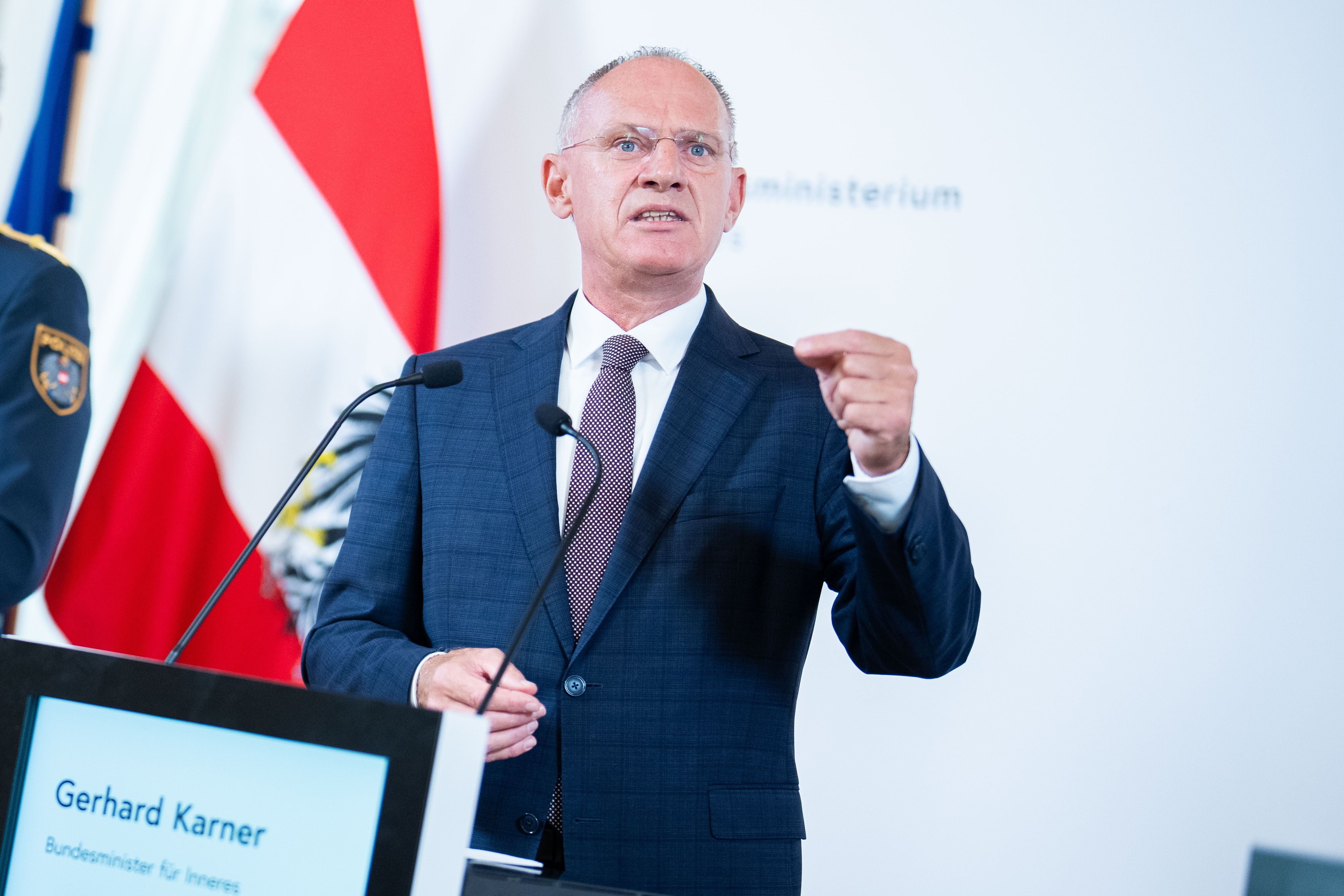 Ein Mann im Anzug spricht bei einer Pressekonferenz vor Mikrofonen und österreichischer Flagge.