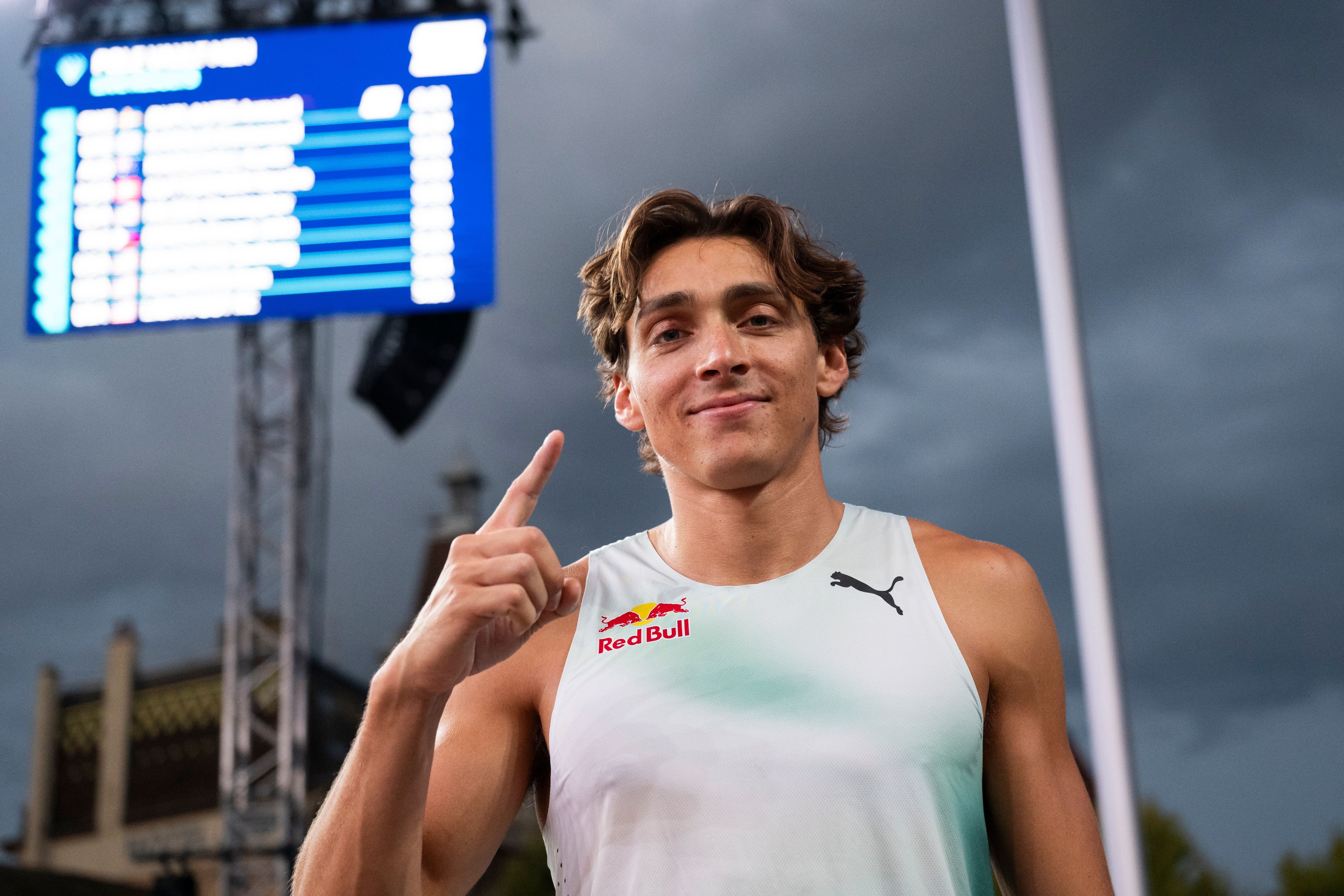 Armand Duplantis blickt lächelnd in die Kamera, deutet mit dem Zeigefinger nach oben und steht vor einer Anzeigetafel im Stadion.