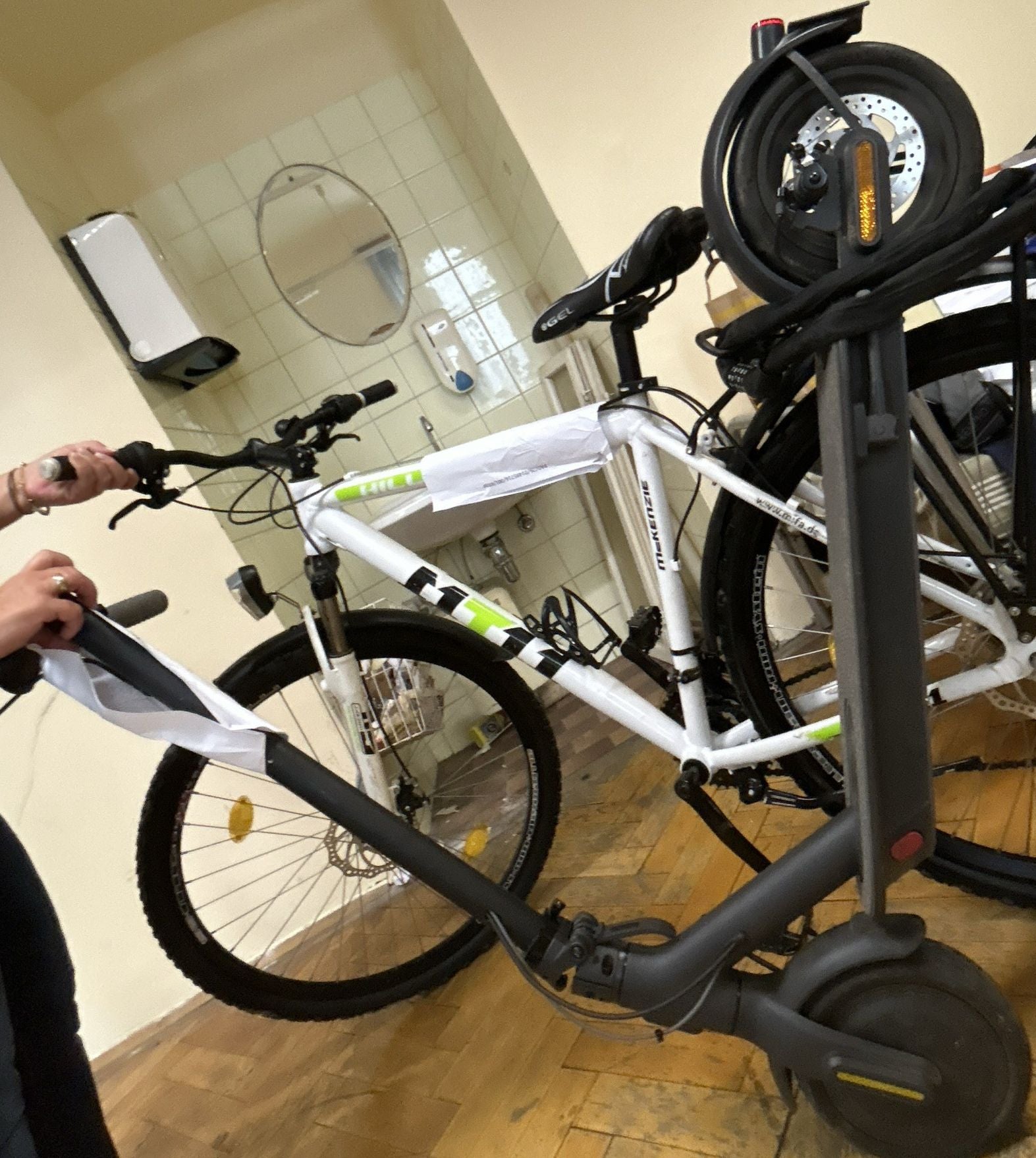Ein Fahrrad und ein E-Scooter stehen nebeneinander in einem Raum mit Waschbecken und Spiegel.
