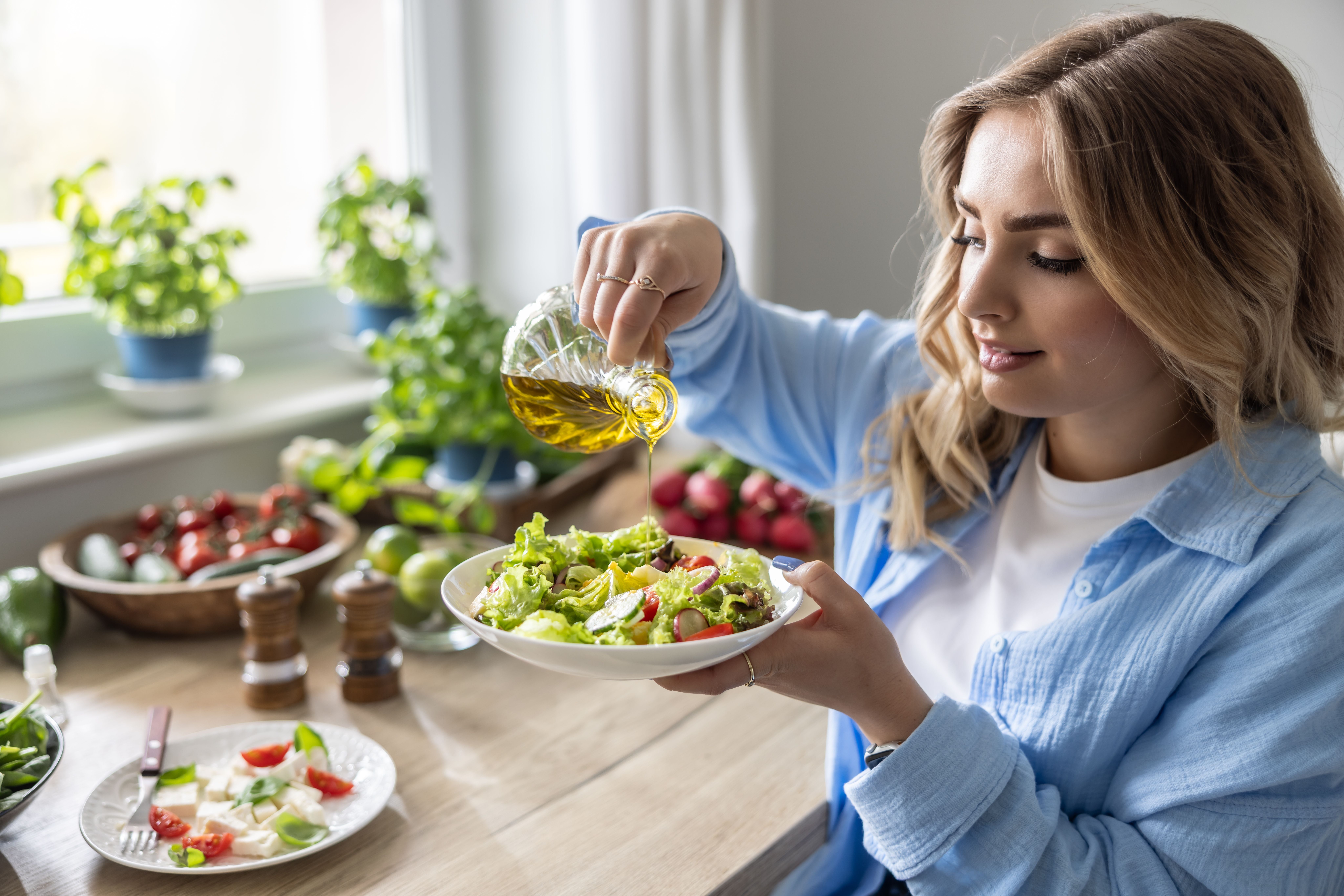 Junge Frau gießt Olivenöl auf einen frischen Salat in einer hellen, gemütlichen Küche.