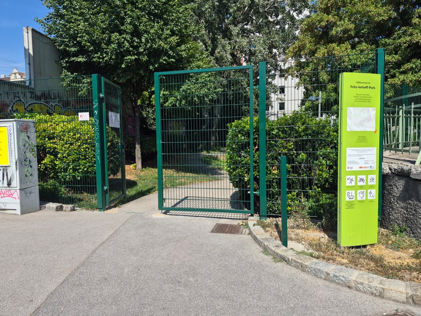 Eingang zum Fritz-Imhoff-Park mit grünem Tor und Hinweistafel bei sonnigem Wetter.