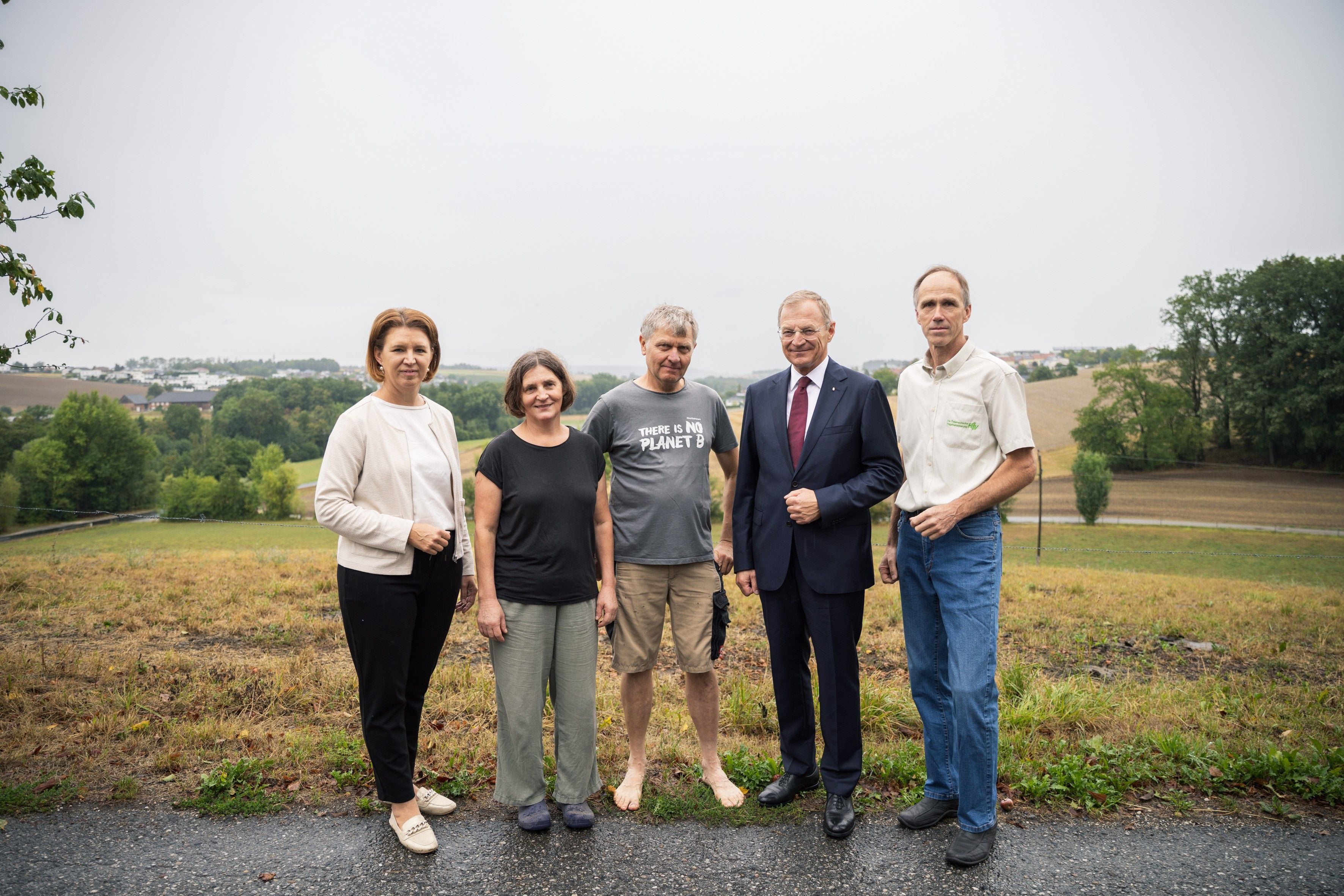Fünf Personen stehen im Freien auf einem Feldweg, im Hintergrund Wiesen und Bäume.
