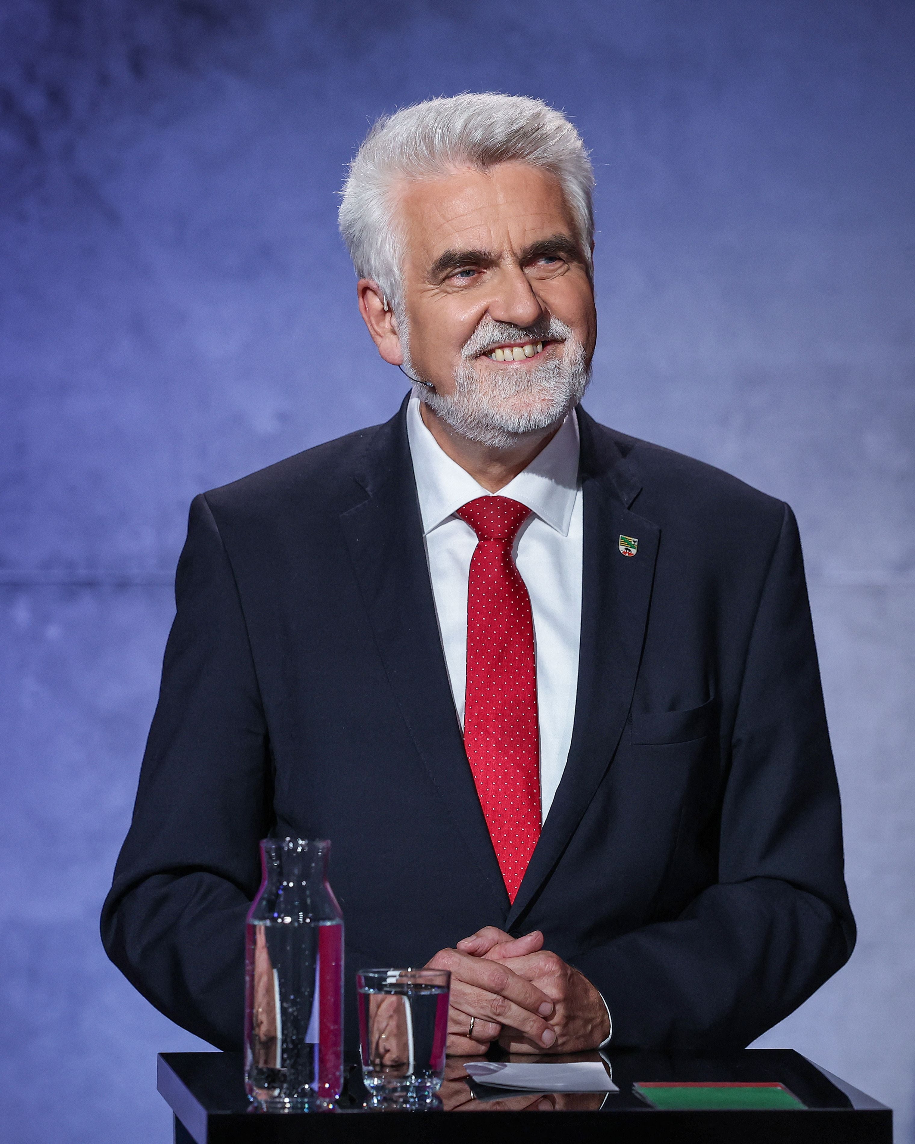 Mann im Anzug mit roter Krawatte steht an einem Pult mit Wasserglas und Karaffe.