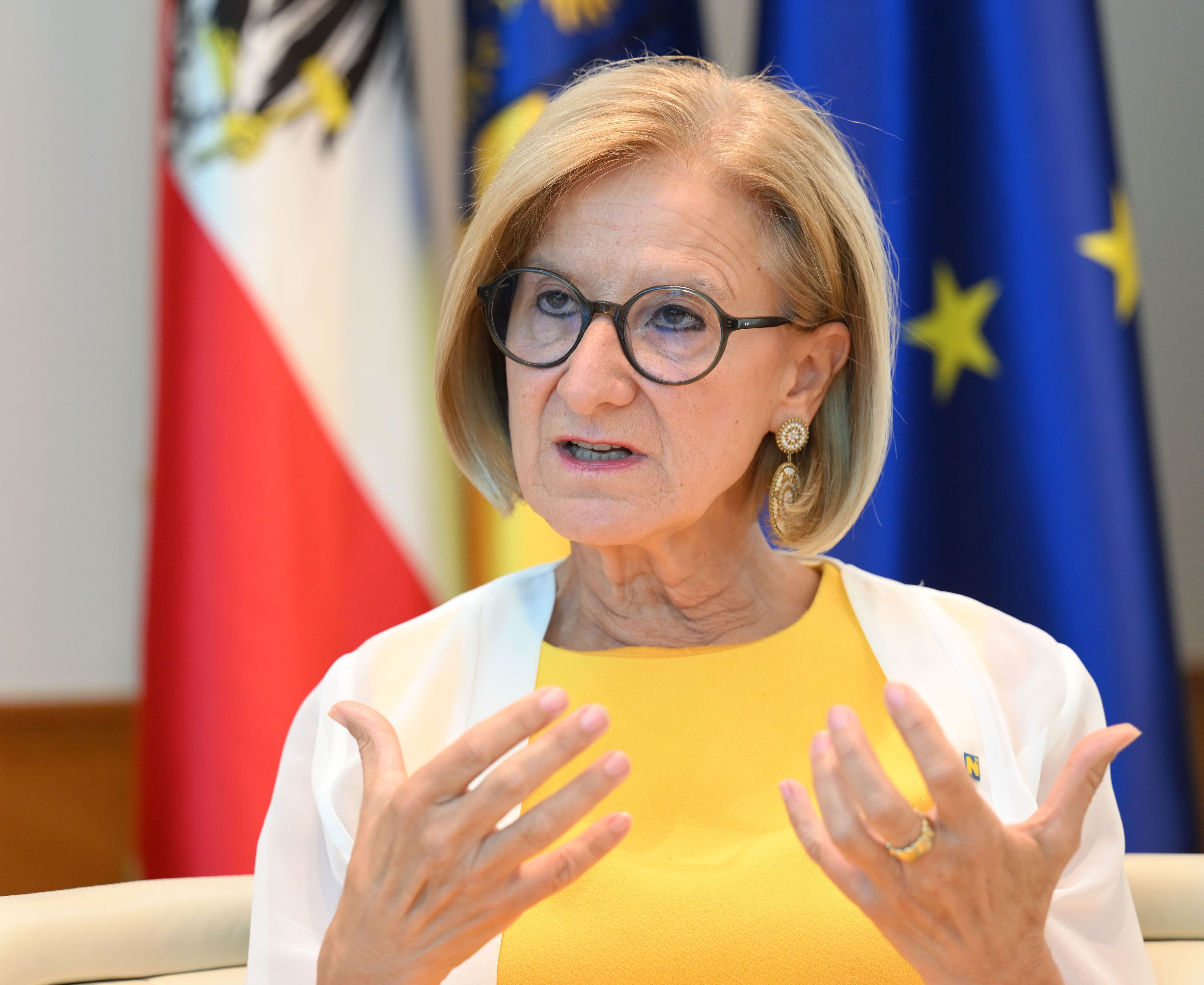 Frau mit Brille spricht gestikulierend vor österreichischer und EU-Flagge.