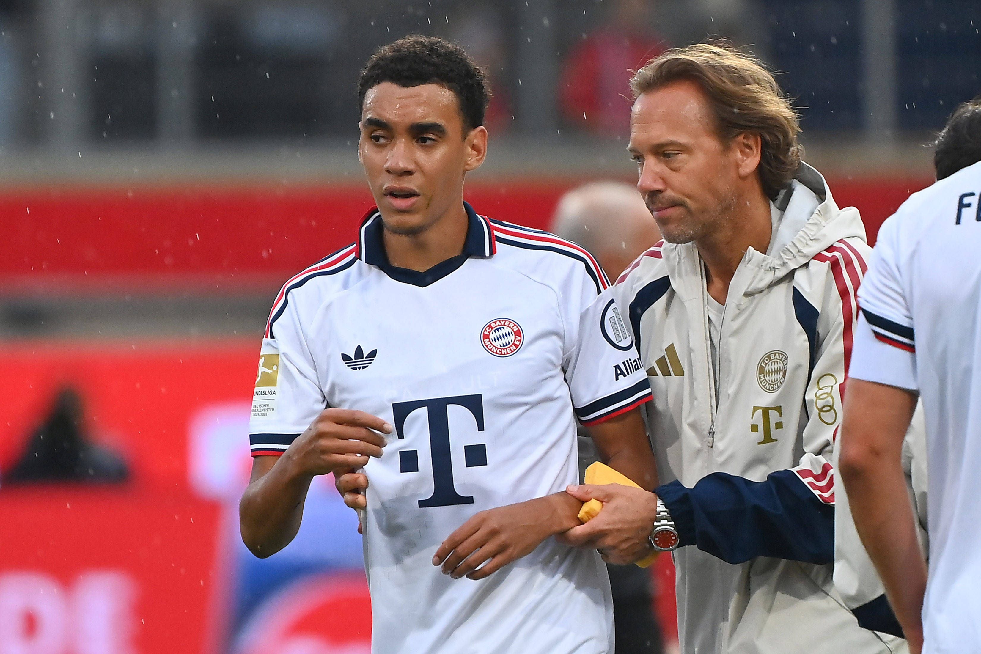 Jamal Musiala wird beim Fußballspiel von Bayern Münchens Teamarzt gestützt vom Platz geführt.
