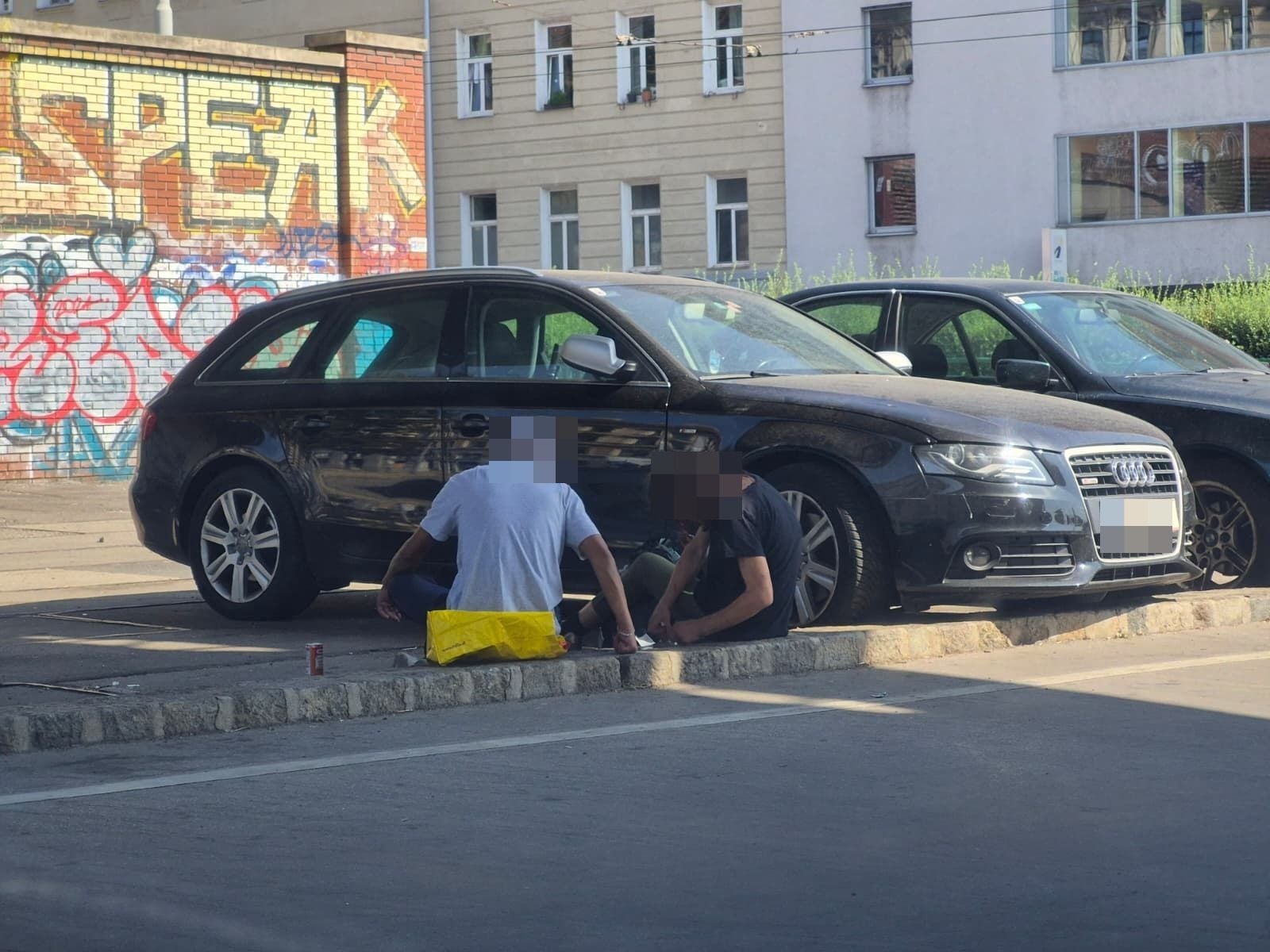 Zwei Personen sitzen vor einem geparkten Auto in einer urbanen Umgebung mit Graffiti an der Wand.