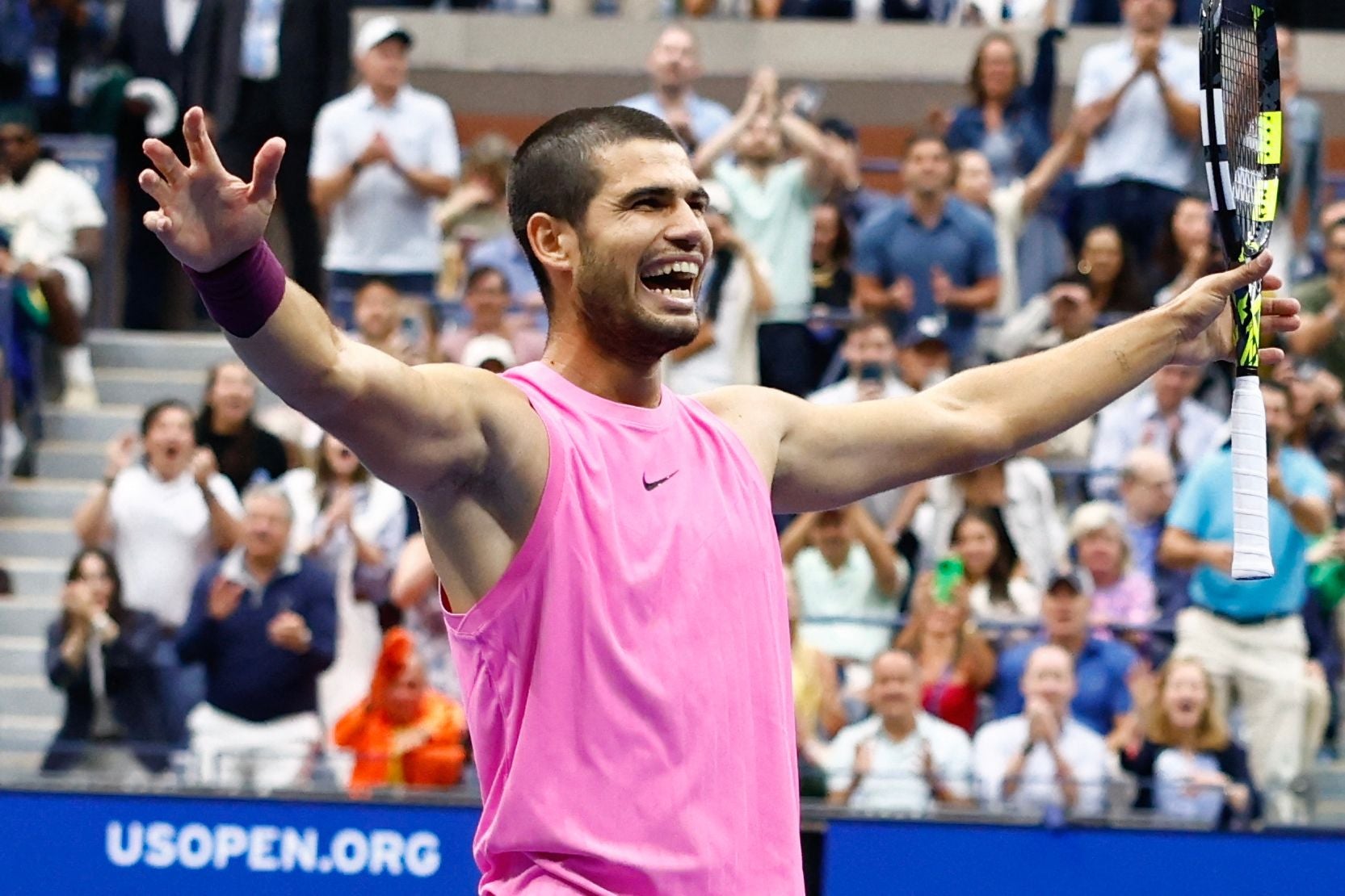 Carlos Alcaraz breitet im rosa ärmellosen Shirt auf dem Tennisplatz strahlend die Arme aus und jubelt dem Publikum zu.