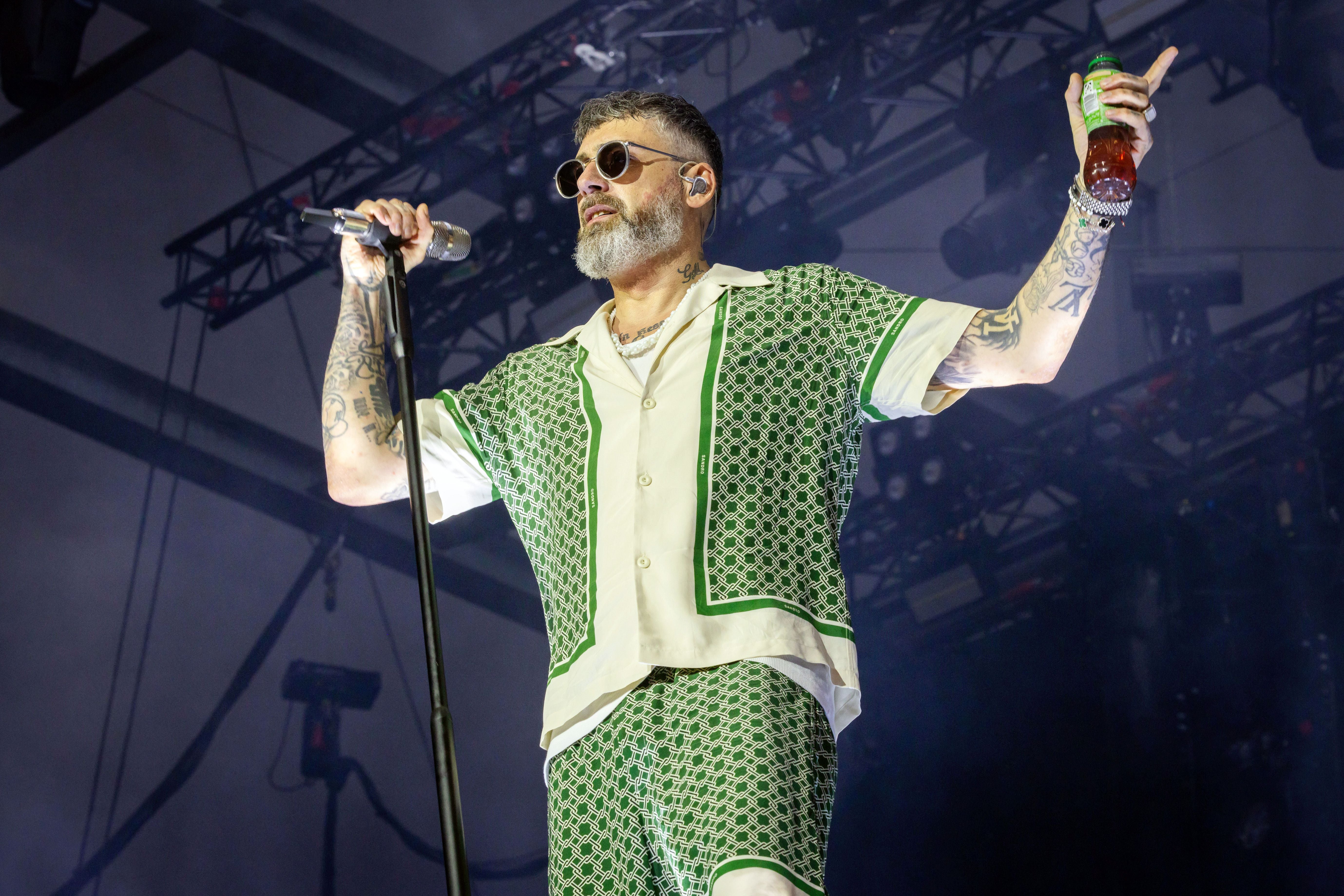 Ein bärtiger Mann mit Sonnenbrille singt auf einer Bühne und hält ein Getränk in der Hand.