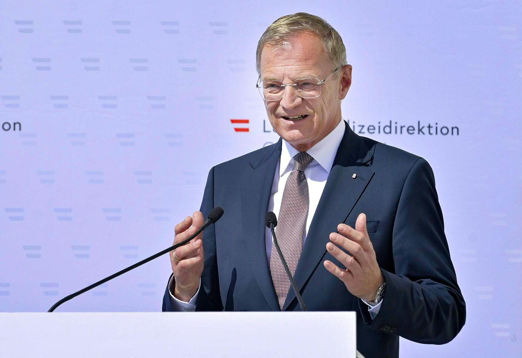 Ein Mann im Anzug spricht an einem Rednerpult mit zwei Mikrofonen vor neutralem Hintergrund.