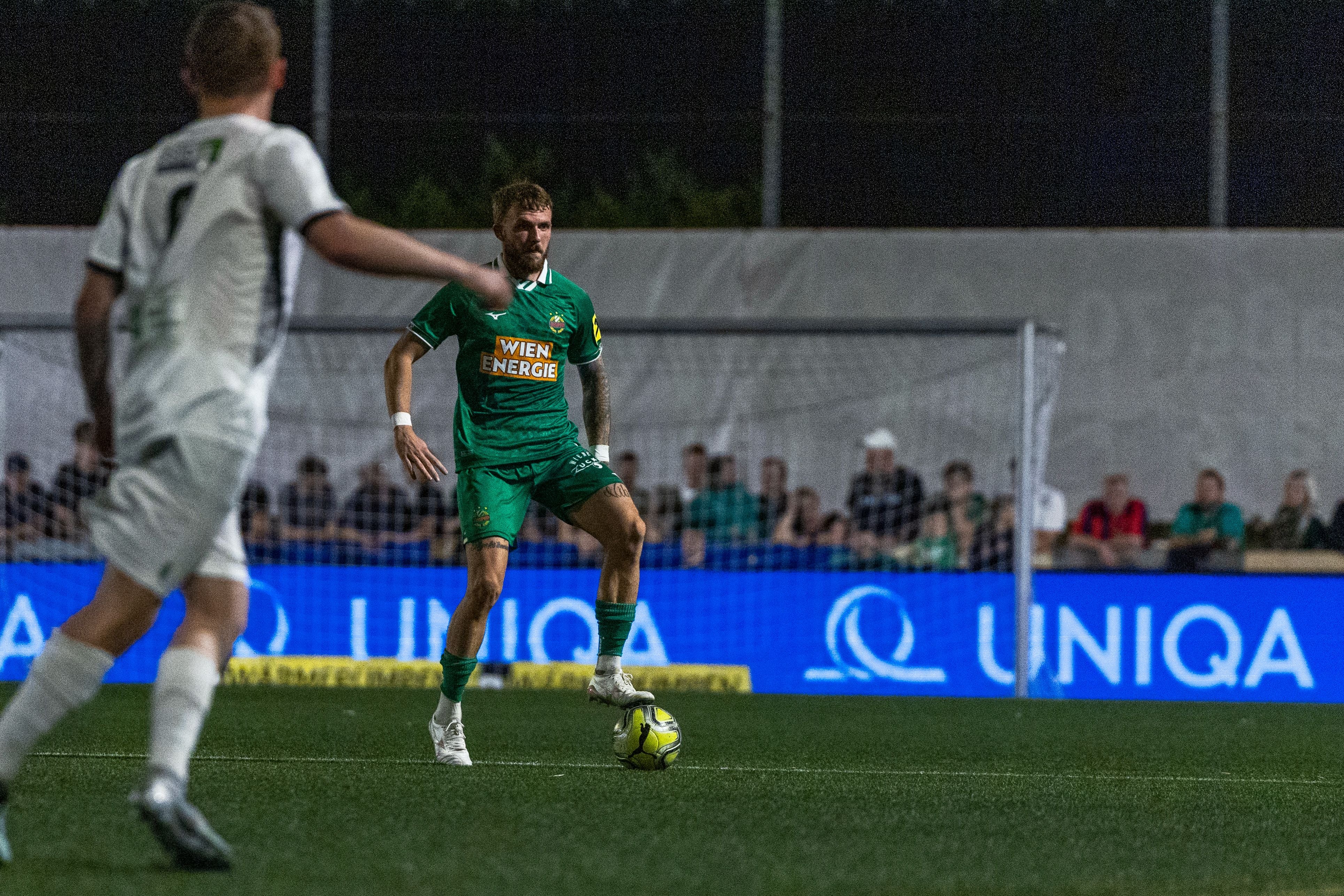 Ein Fußballspieler von Rapid Wien steht mit Ball am Fuß auf dem Spielfeld, Zuschauer im Hintergrund.