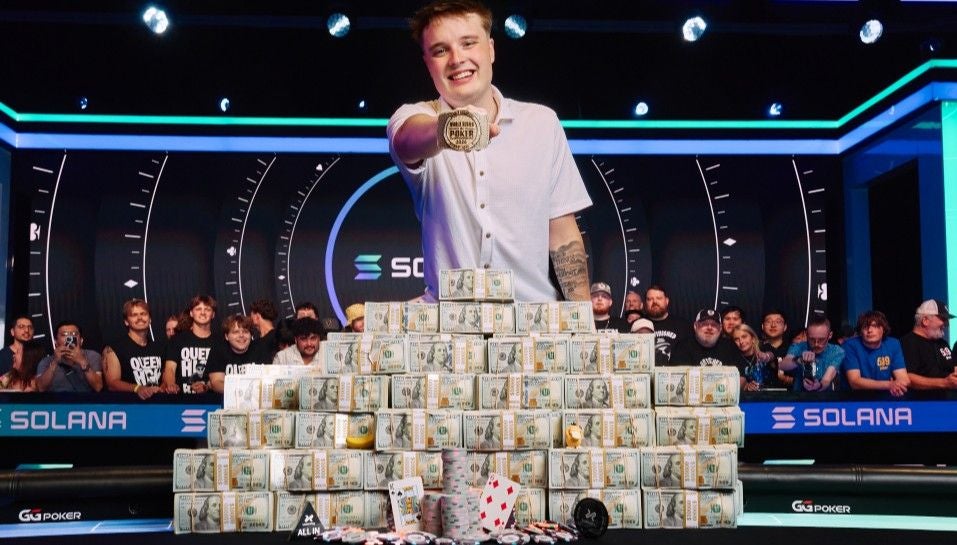 Ein junger Mann steht vor einem Berg aus Geldbündeln und hält stolz eine Poker-Gewinnermedaille hoch.