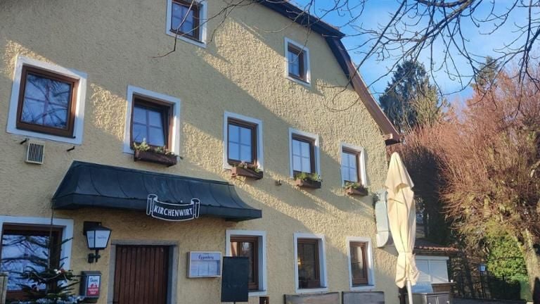 Gelbes Gasthaus mit weißen Fensterrahmen und Sonnenschirm vor dem Eingang bei Tageslicht.