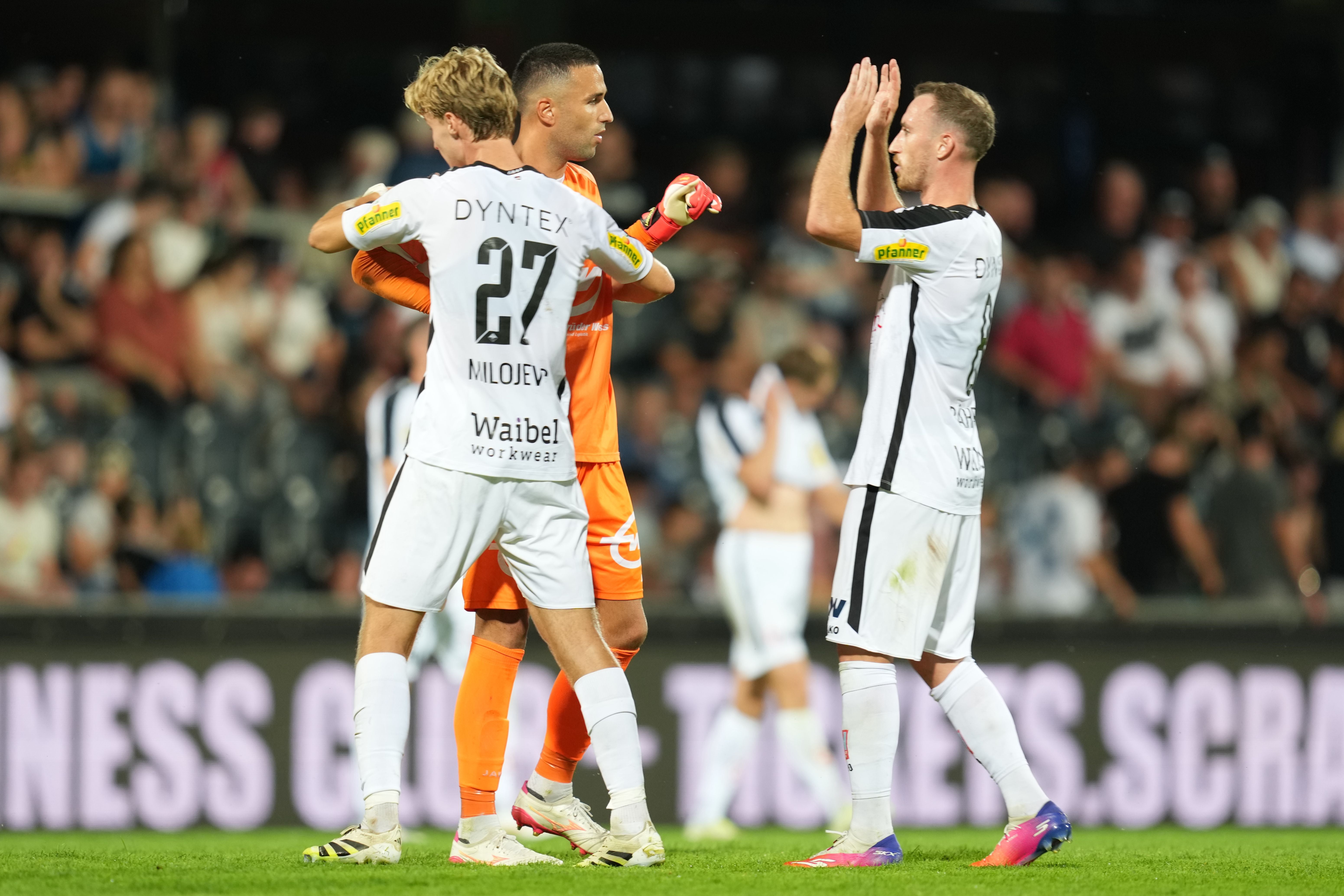 Ein Fußballspieler mit der Rückennummer 27 im weißen Trikot umarmt einen Torwart im orangen Dress, während ein anderer Spieler daneben klatscht.