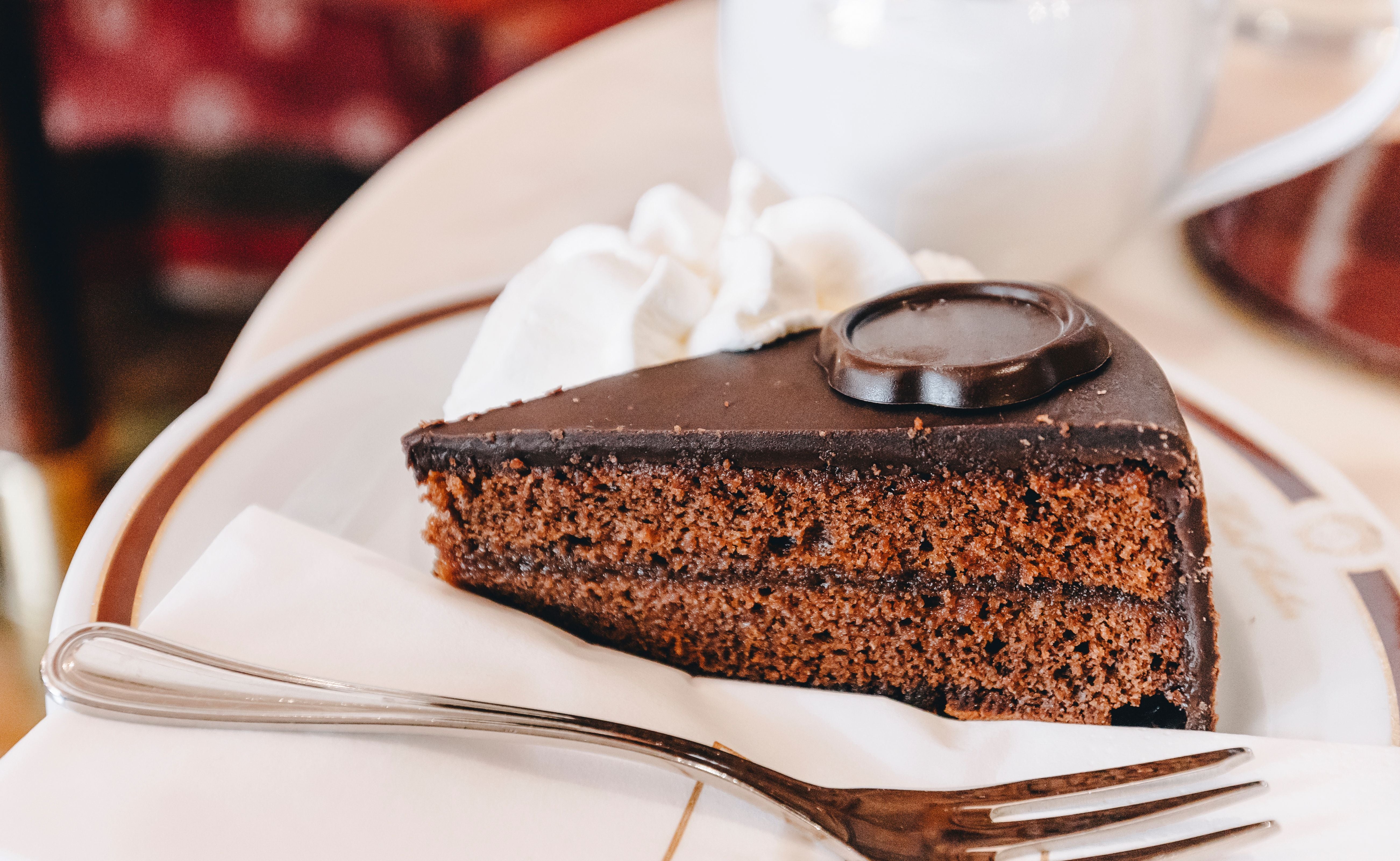 Ein Stück Sachertorte mit Schokoladenglasur und Sahne auf einem weißen Teller mit Gabel.