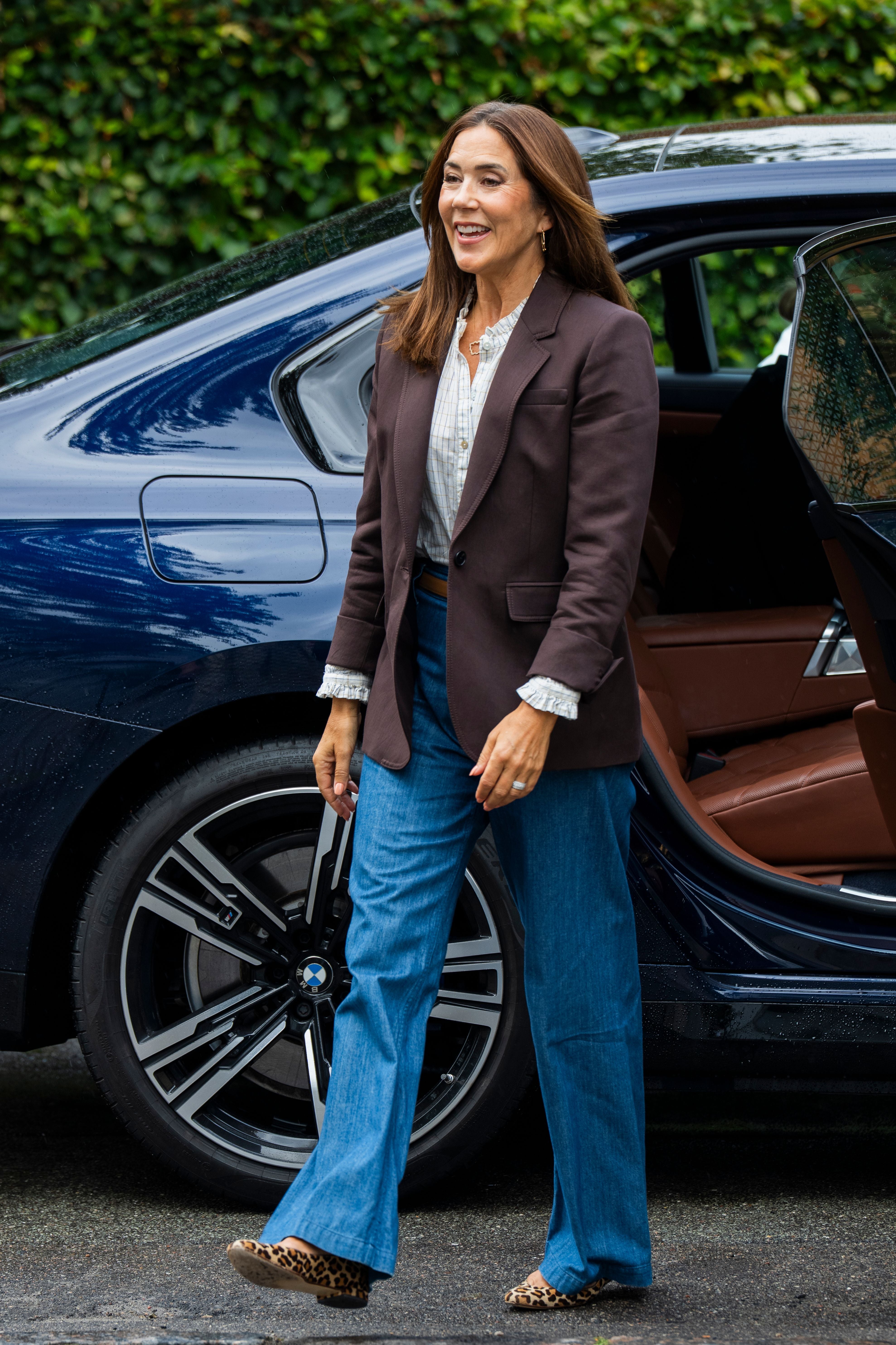 Eine Frau steigt lächelnd aus einem Auto und trägt Blazer und Jeans.