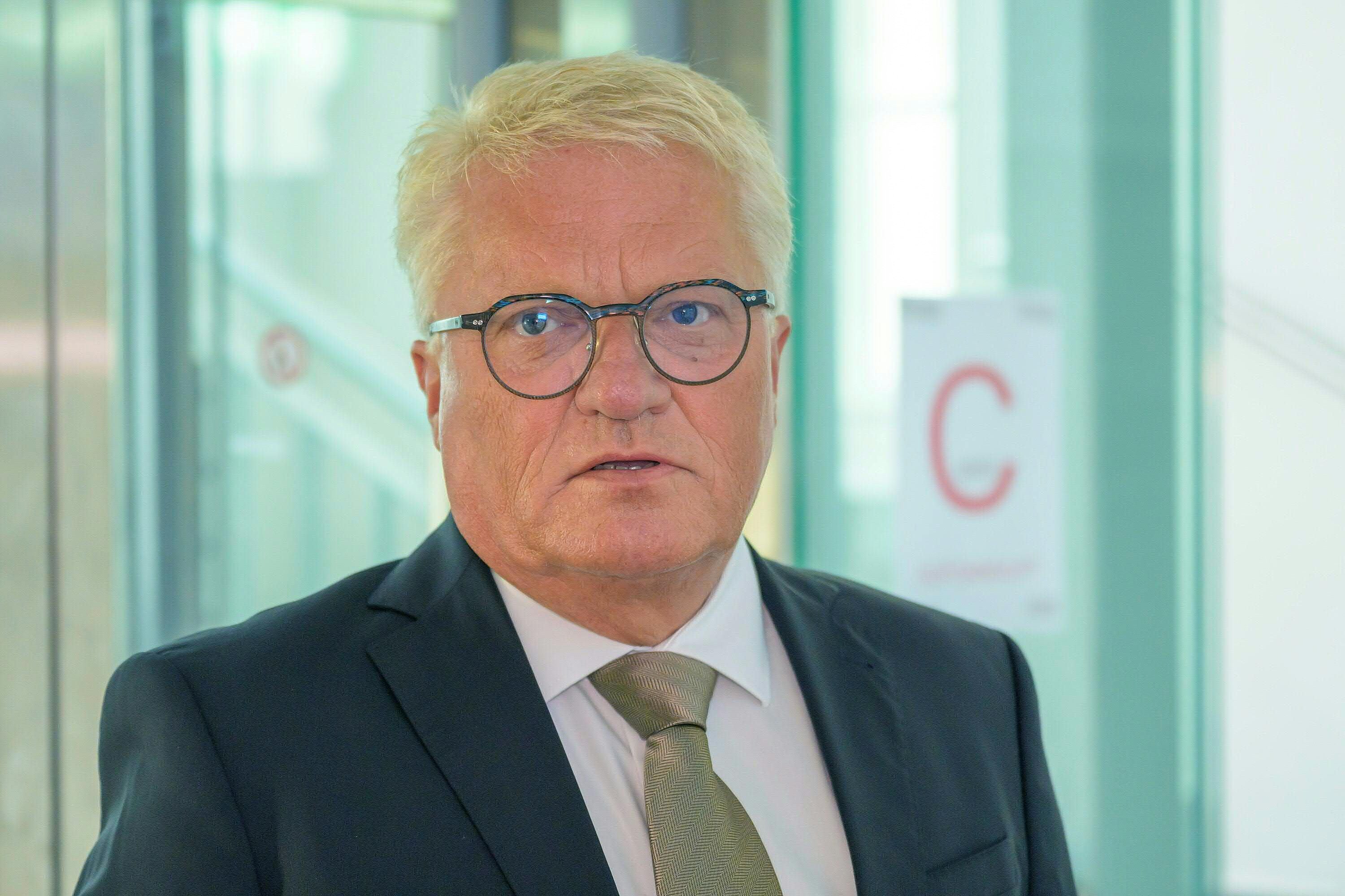 Ein Mann im Anzug mit Brille steht in einem modernen Gebäude und blickt ernst in die Kamera.