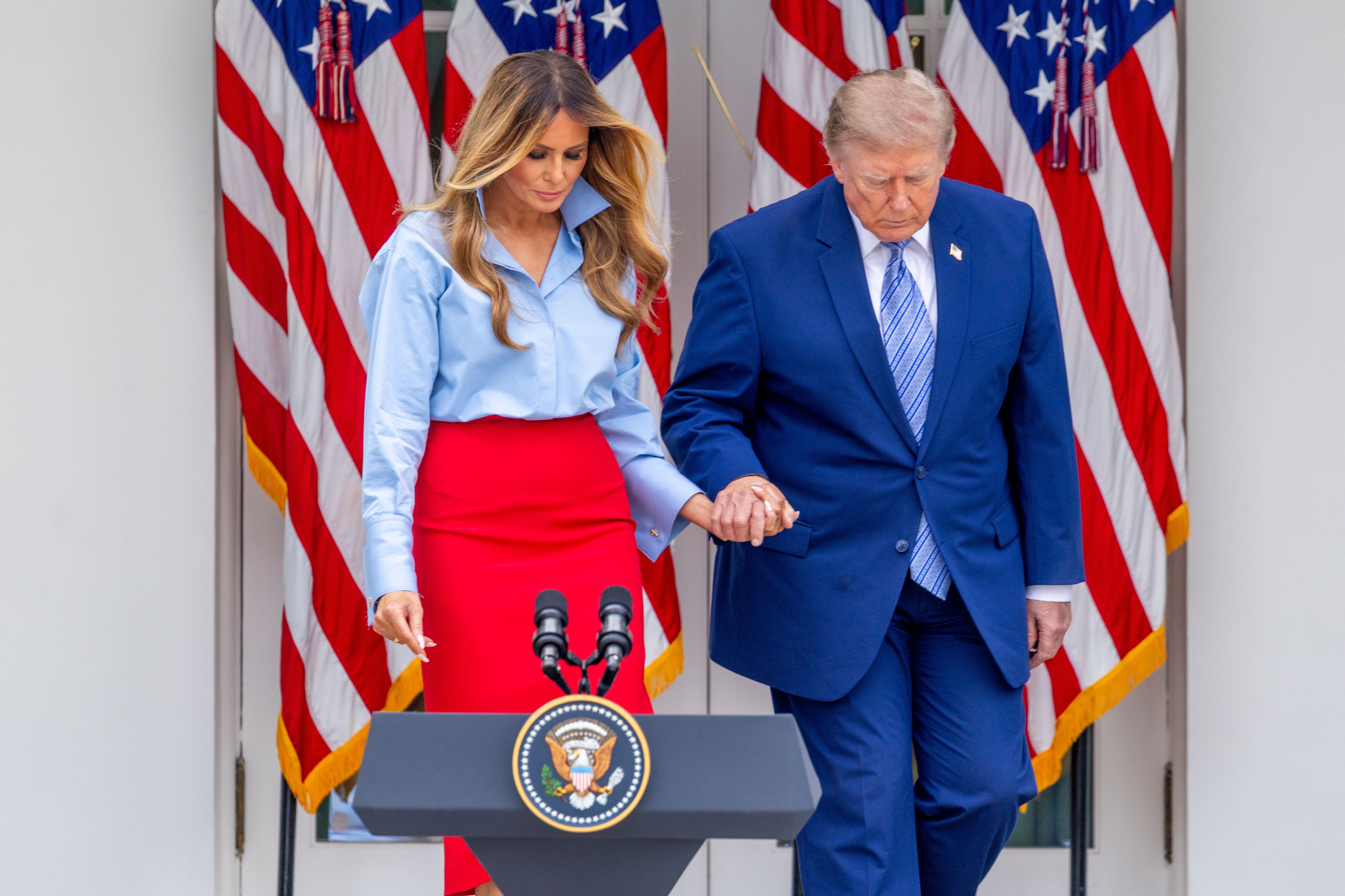Melania Trump und Donald Trump gehen Hand in Hand im Rosengarten vor US-Flaggen.