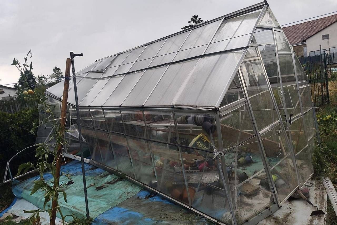 Ein umgekipptes Gewächshaus liegt schief im Garten, umgeben von Gartenwerkzeug und Pflanzen.