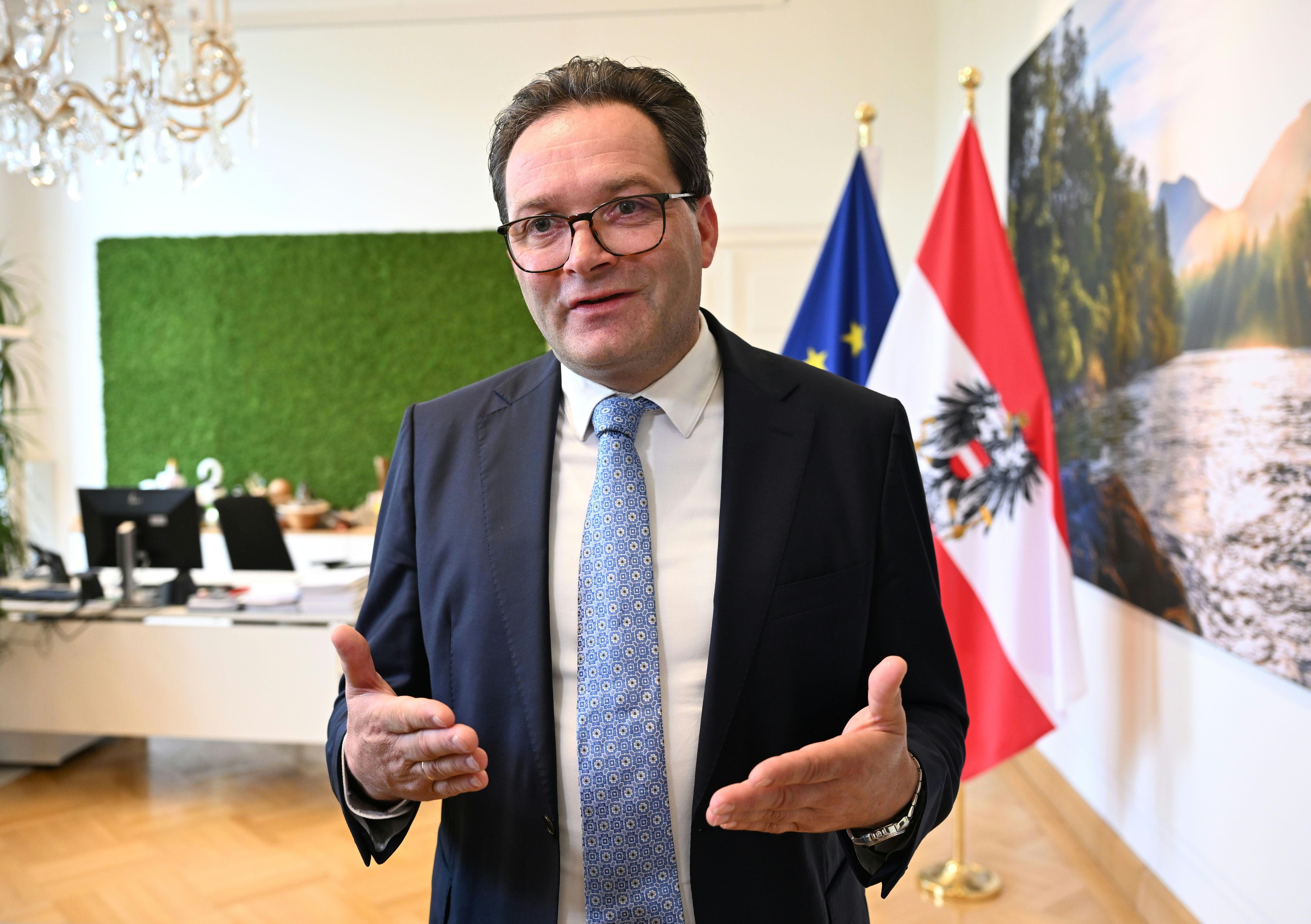 Ein Mann im Anzug spricht in einem Büro, im Hintergrund sind EU- und Österreich-Flaggen.