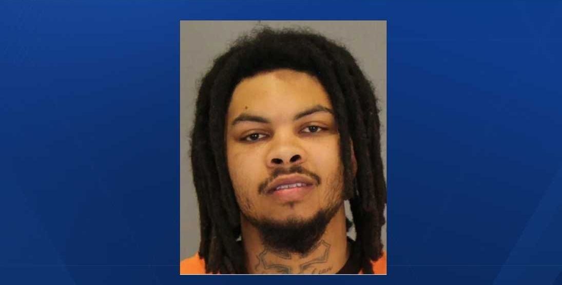 Porträt eines jungen Mannes mit Dreadlocks und Hals-Tattoo vor neutralem Hintergrund.