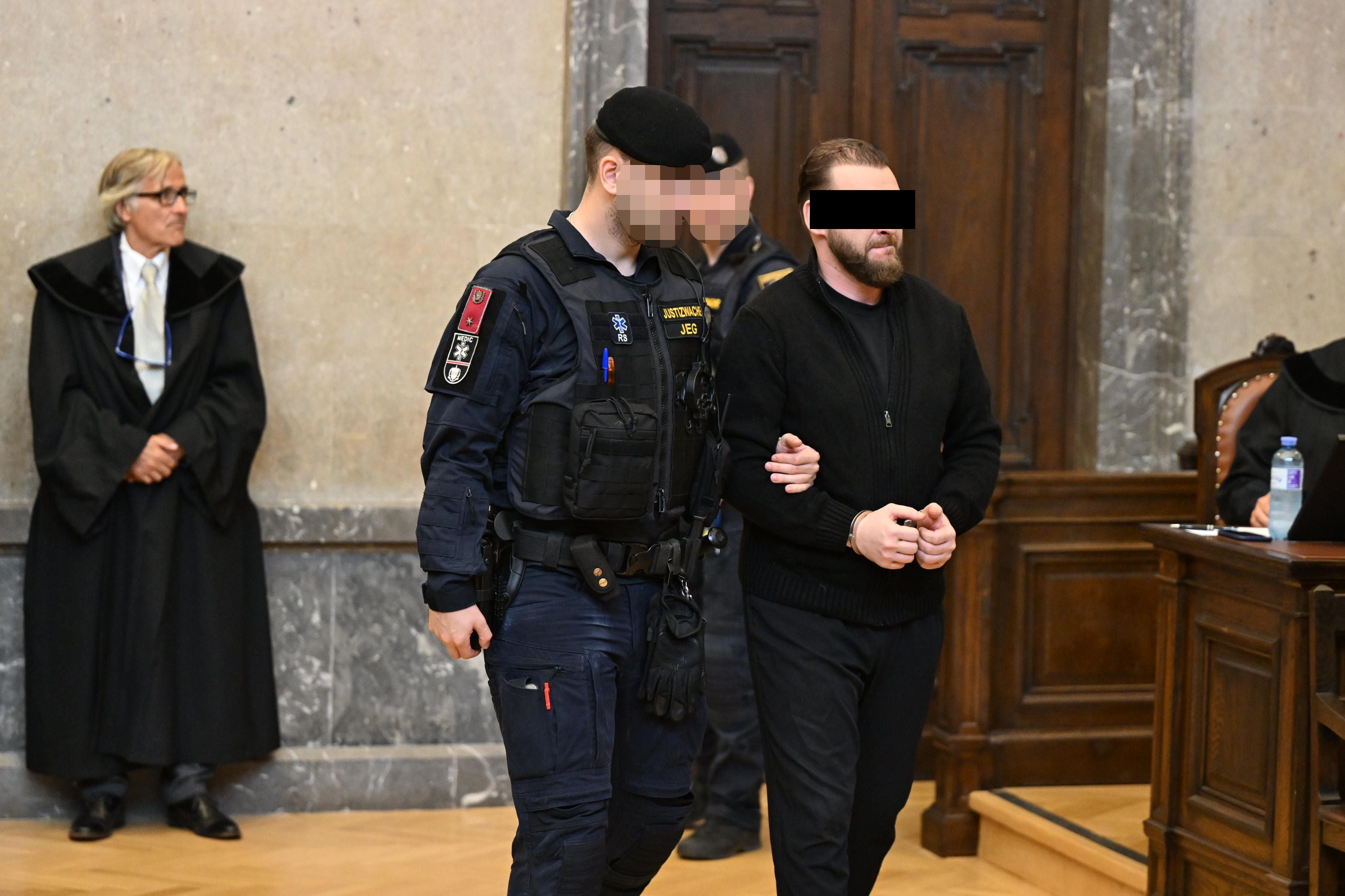 Ein Polizist führt einen Angeklagten in Handschellen vor Gericht, weitere Personen im Hintergrund.
