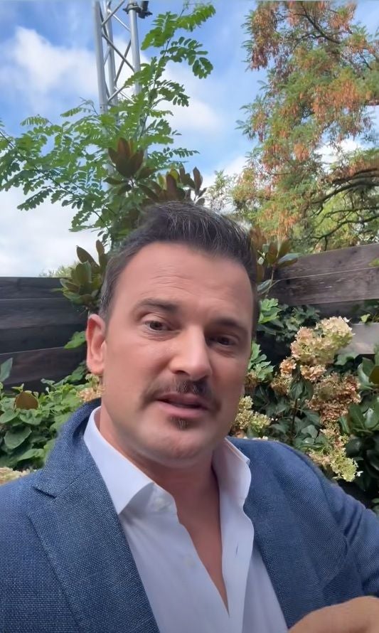 Ein Mann im blauen Sakko steht draußen vor grünen Sträuchern und Blumen.