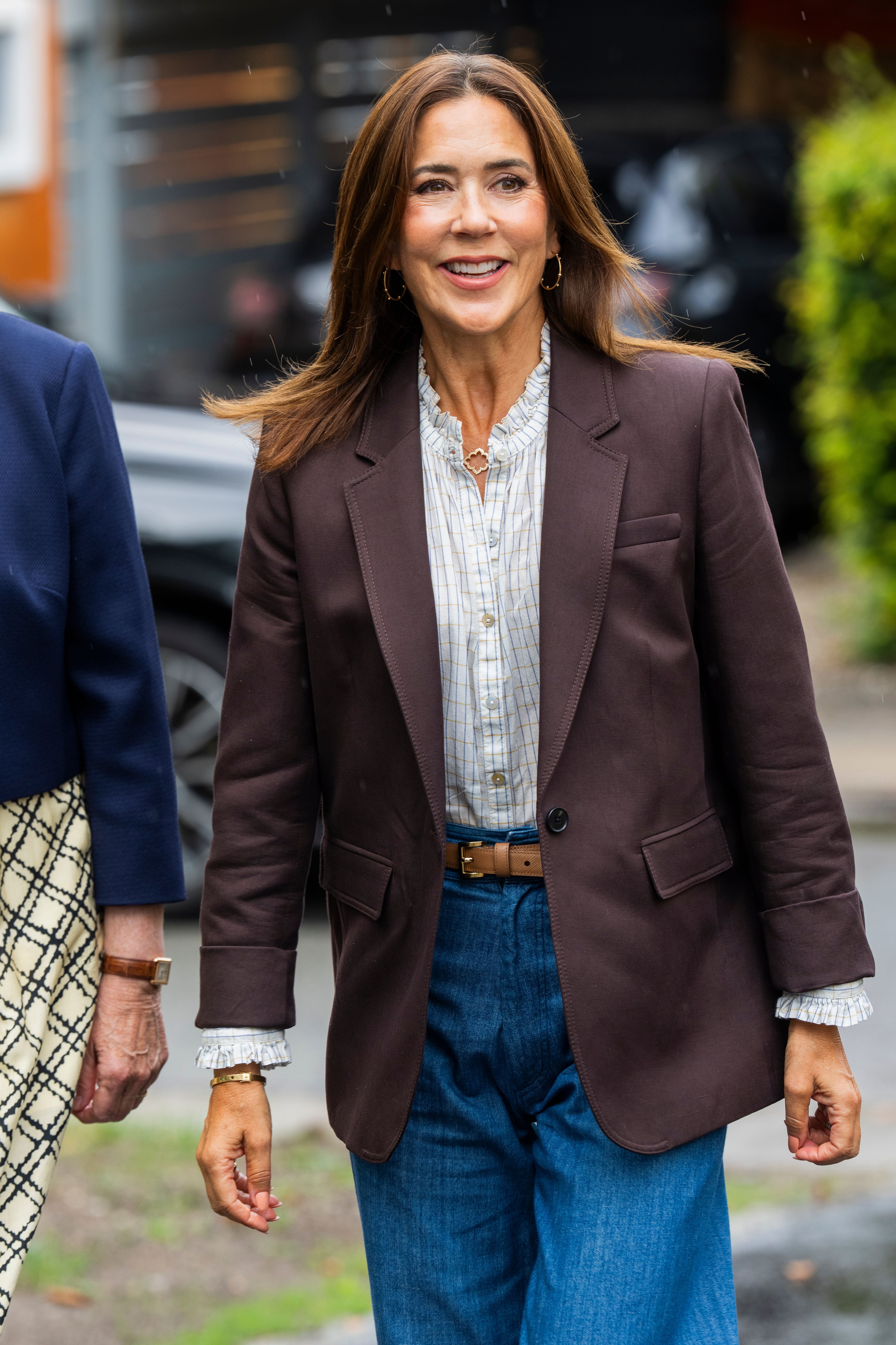 Frau mit braunem Blazer und blauer Jeans lächelt im Freien.
