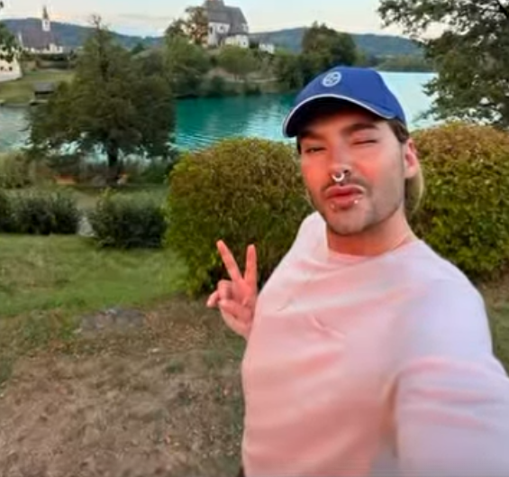 Person mit Basecap macht ein Selfie mit Friedenszeichen vor einem See und einer Kirche im Hintergrund.