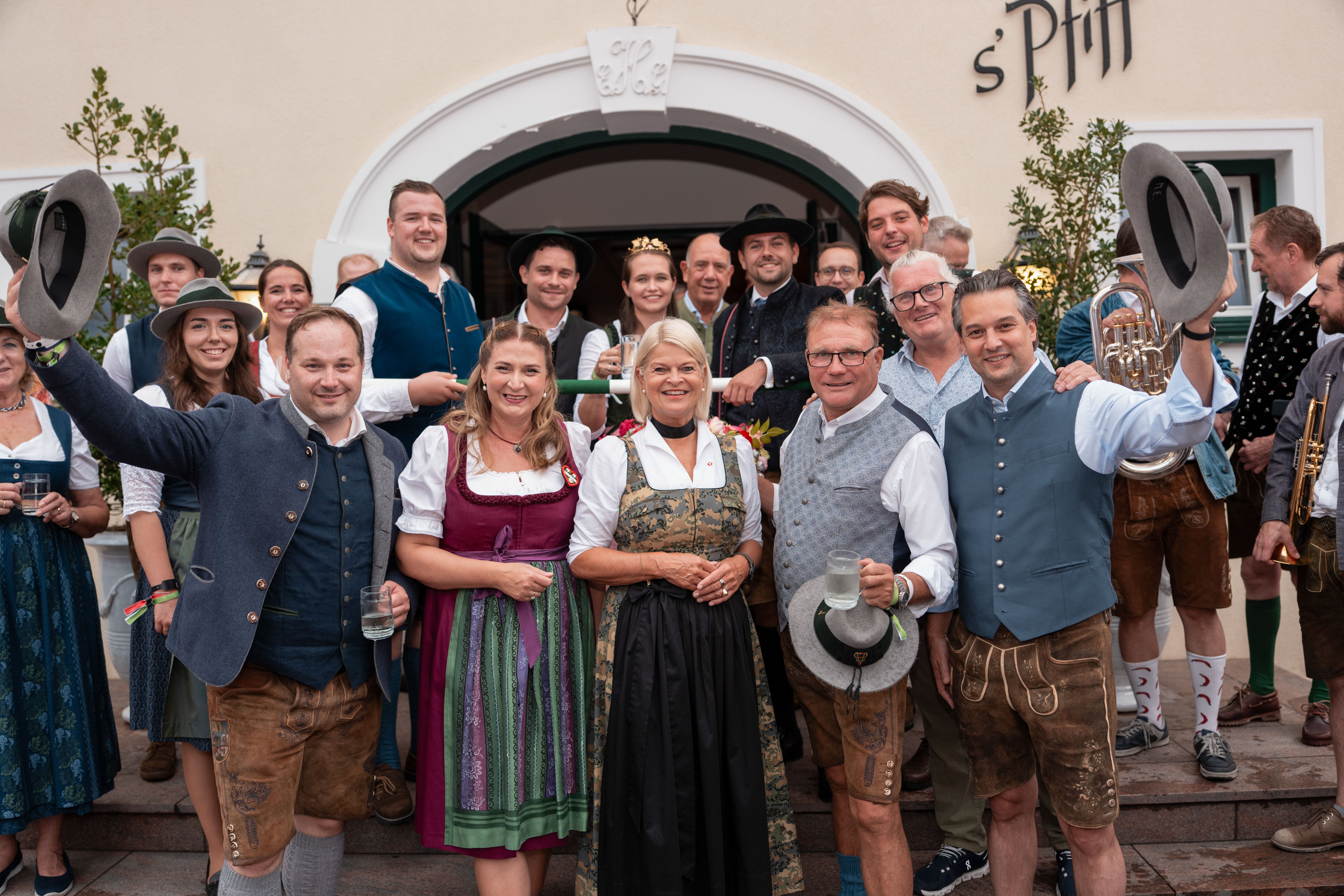 Gruppe in traditioneller Tracht feiert fröhlich vor einem Gebäude, einige halten Krüge und Hüte hoch.